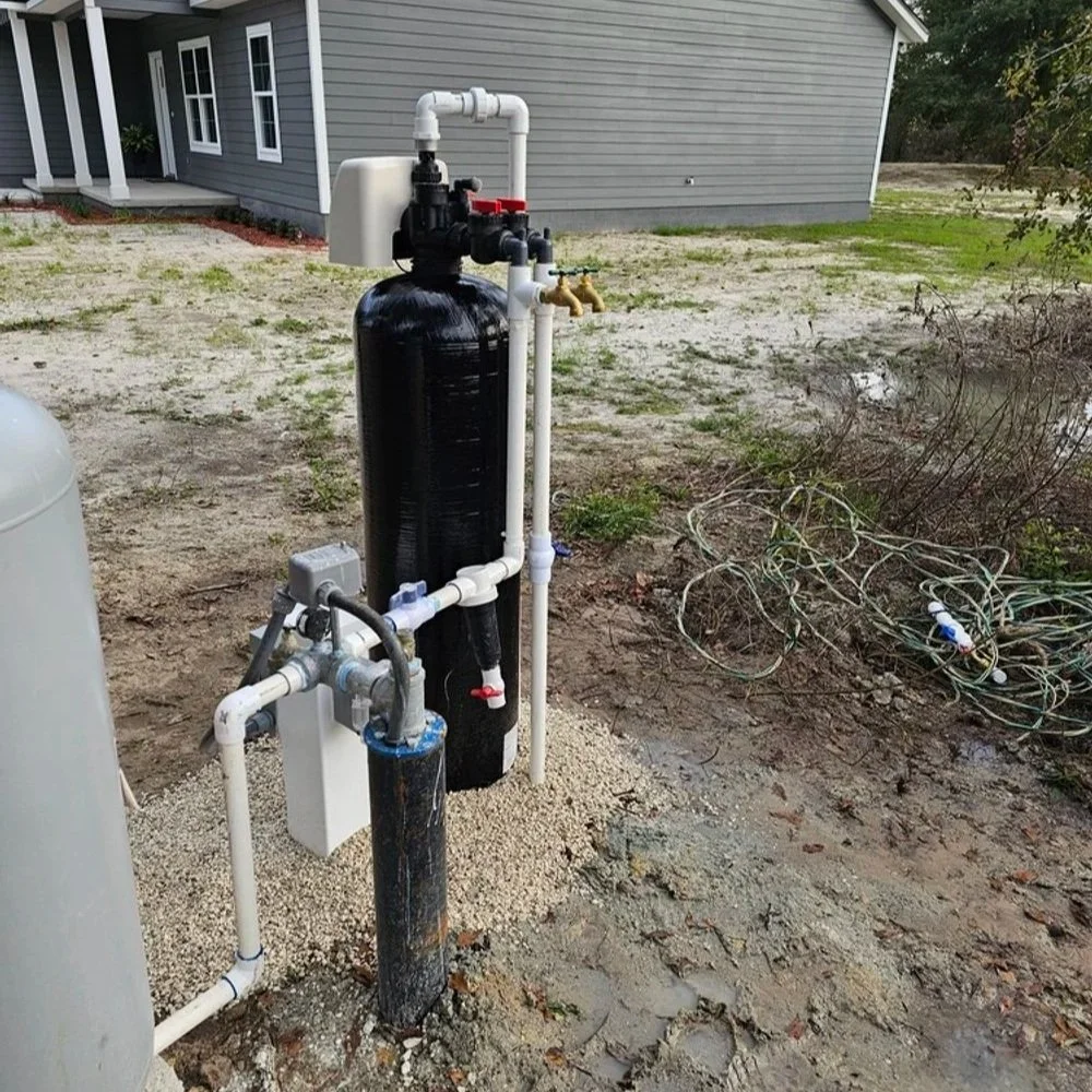 Outdoor water filtration system next to a house with a gray exterior, showing a large black filter tank with white pipes, valves, and a smaller filter, on a gravel surface amid dirt and some wire.