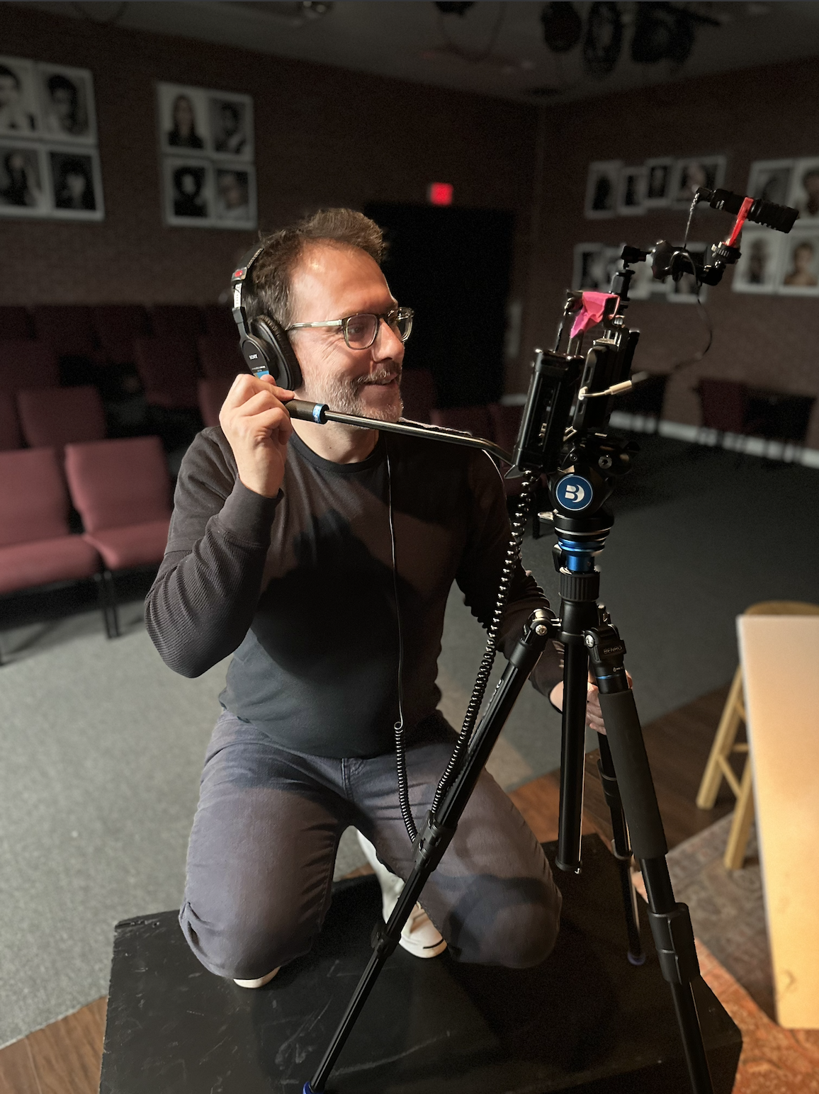 A man wearing glasses and headphones, smiling and kneeling in front of a camera on a tripod in a dimly lit room with theater seats and framed photos on the wall.