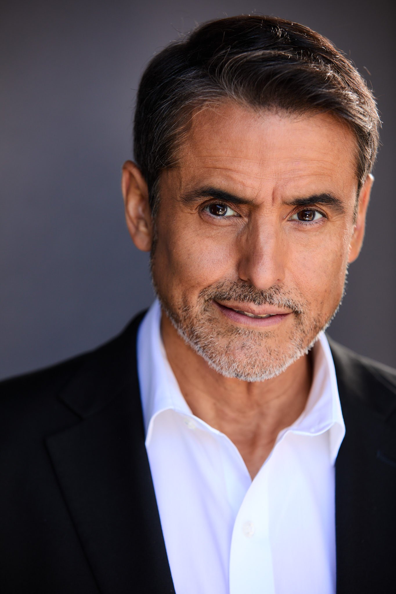 Close-up of a middle-aged man with dark hair streaked with gray, wearing a white shirt and black blazer, looking directly at the camera with a slight smile.