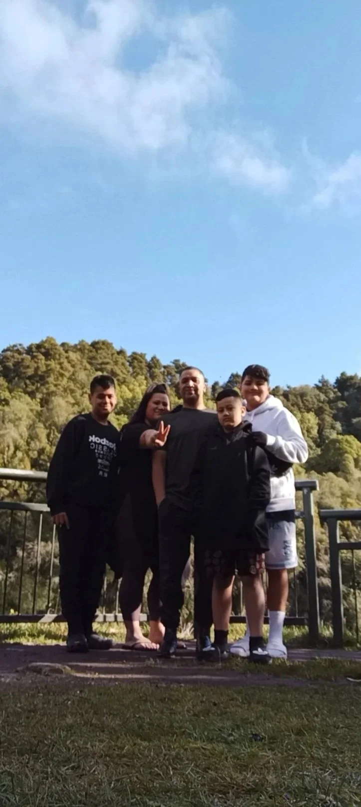 Group of five people standing outdoors on a wooden platform with a forested hillside in the background, under a partly cloudy sky.