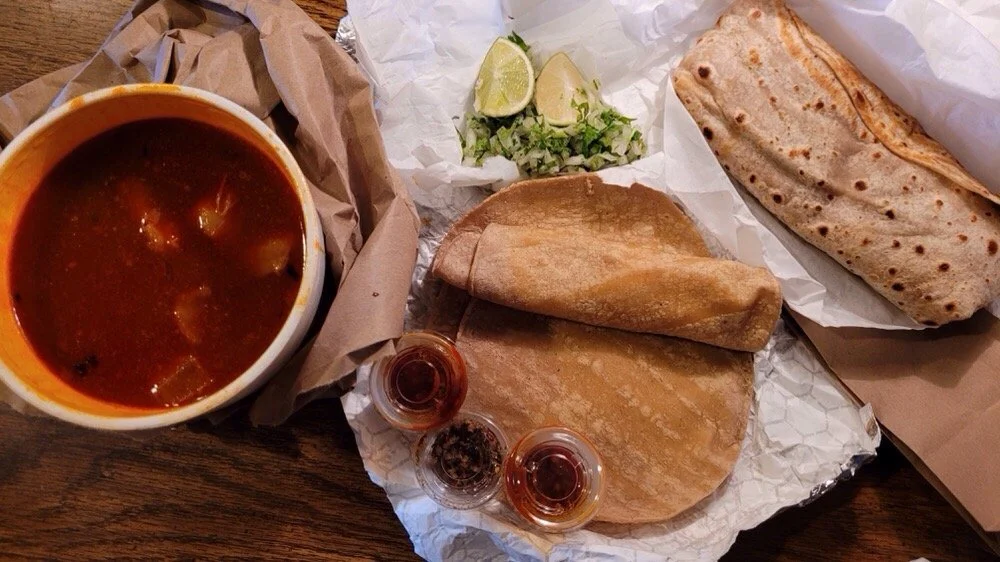 Mexican meal with a bowl of chili, two tacos with pita bread, lime wedges, chopped onions, and small containers of hot sauce on a wooden table.