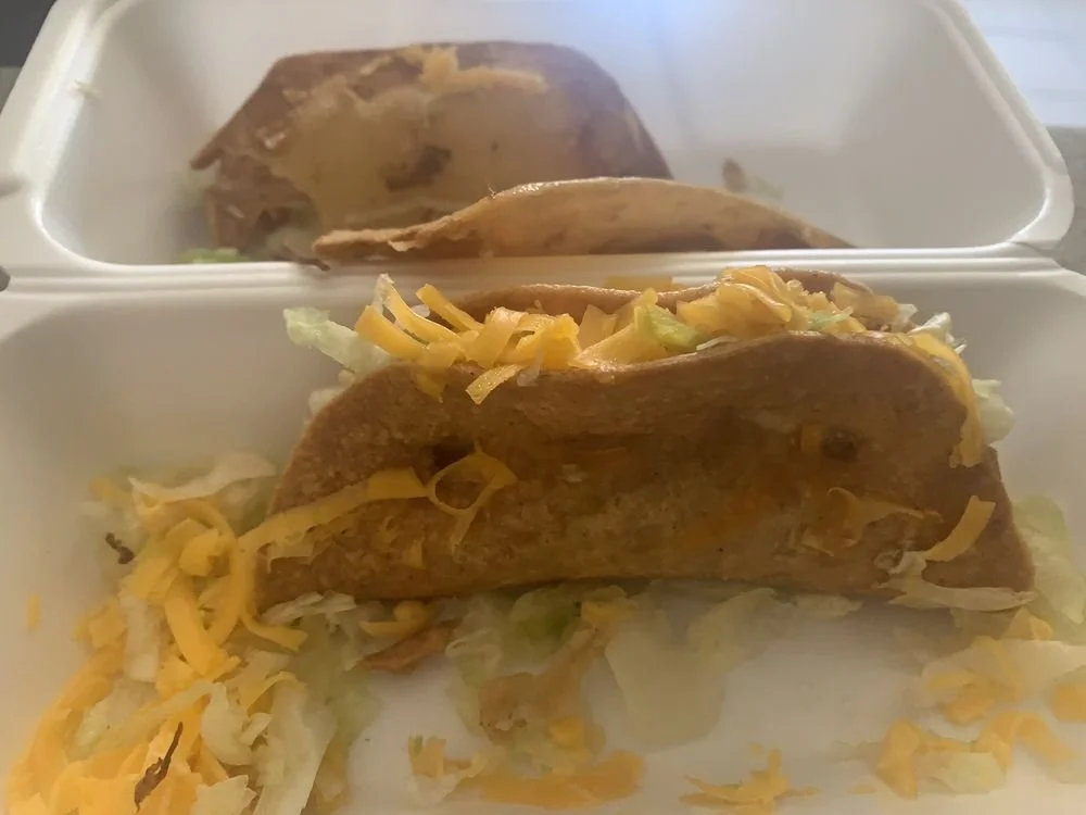 Two tacos with shredded cheese, lettuce, and taco meat in a white takeout container.