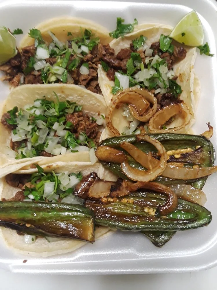 Three street-style tacos filled with chopped meat and topped with diced onions and cilantro, served with grilled jalapeños and lime wedges.