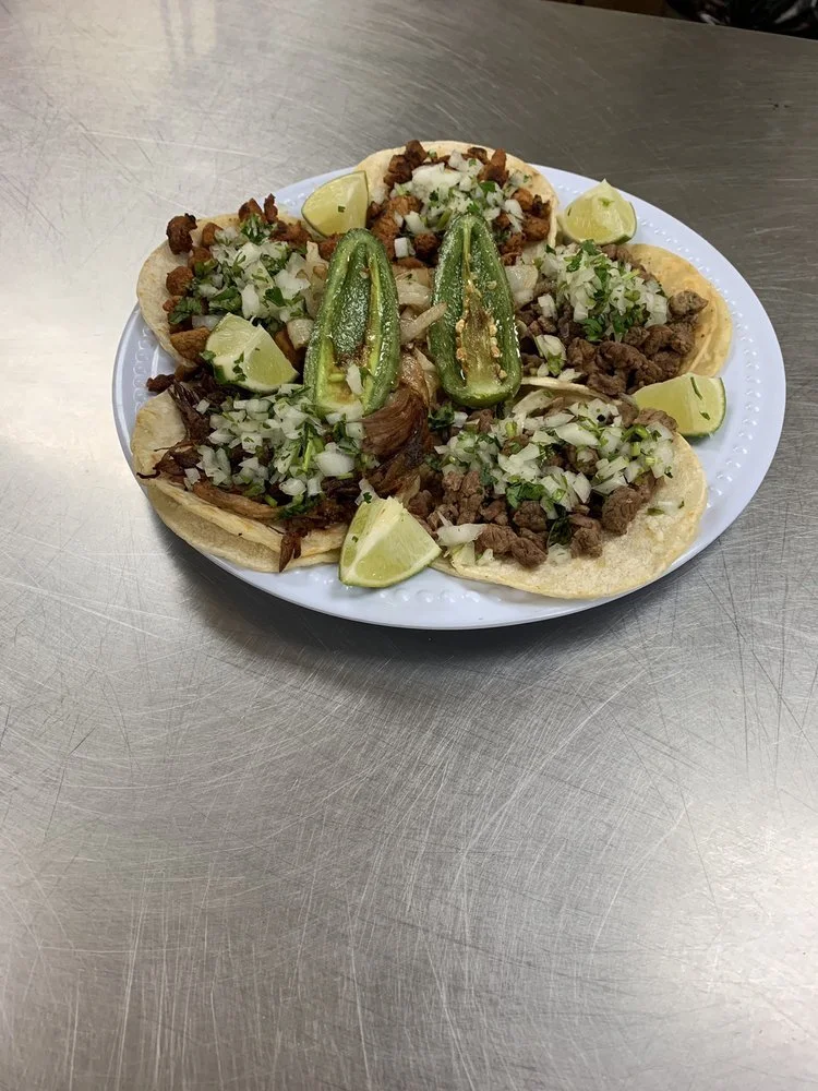 Plate of five tacos with chopped onions, cilantro, grilled meat, and sliced jalapeños, garnished with lime wedges.