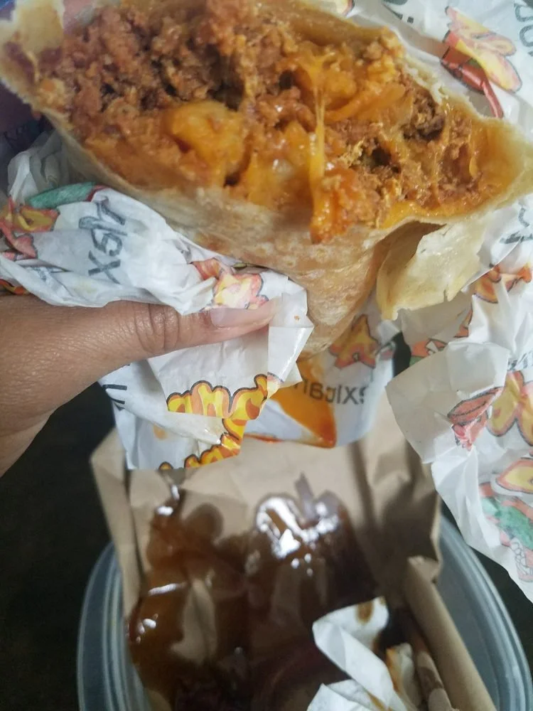 Close-up of a person holding a large beef and cheese burrito wrapped in branded paper, with some sauce visible on the paper, and a small container of dipping sauce nearby.