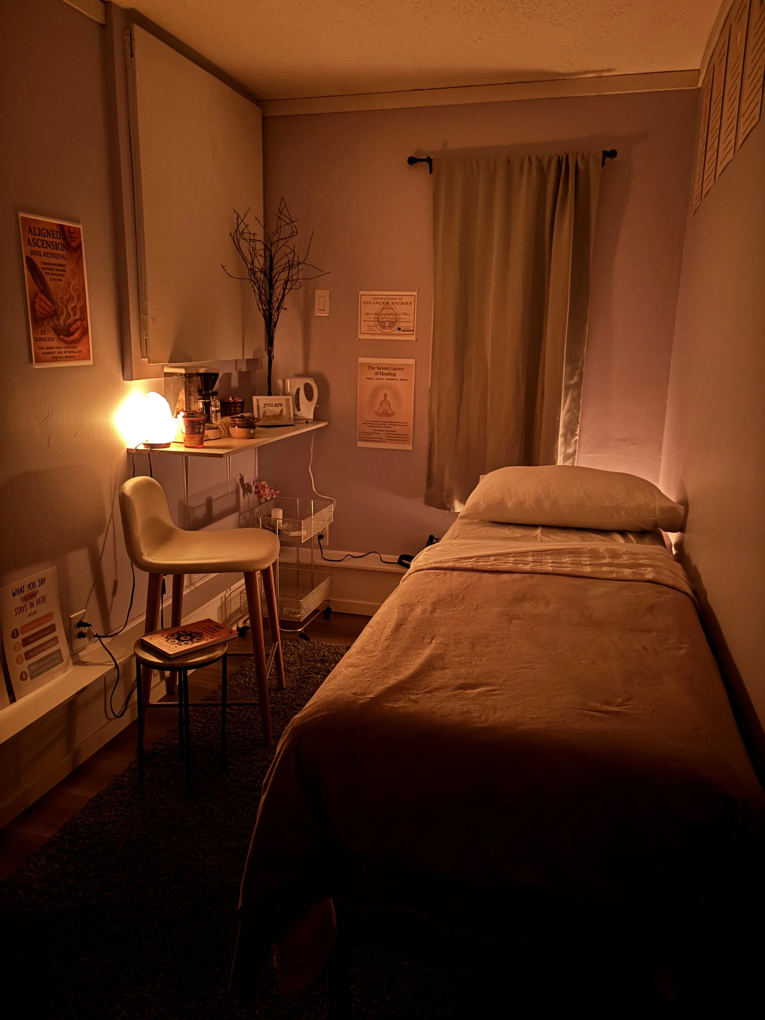 A dimly lit bedroom with a bed on the right, a window with curtains, a small table with a chair, lamps, and framed posters on the walls.