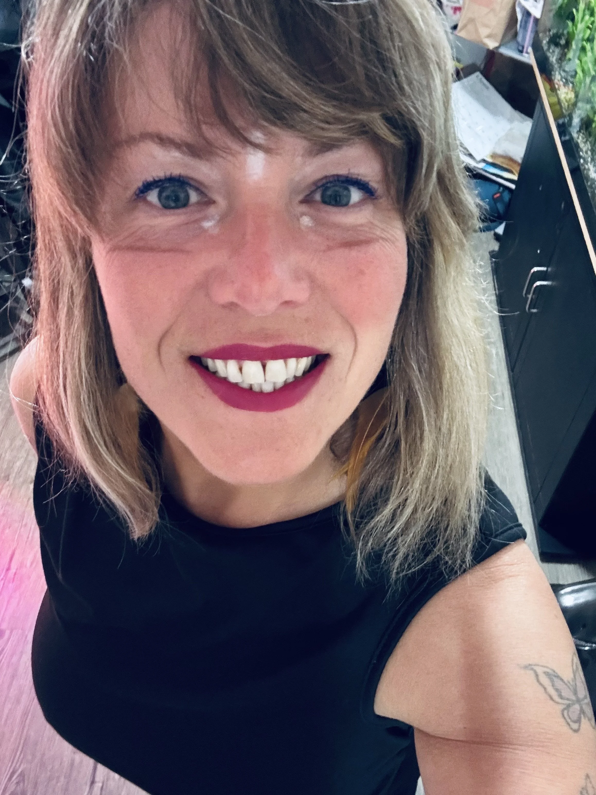 Close-up of a smiling woman with blue eyes, light makeup, and shoulder-length light brown hair, wearing a black sleeveless top and showing a tattoo of a butterfly on her upper arm, in an indoor setting with a desk and plants in the background.