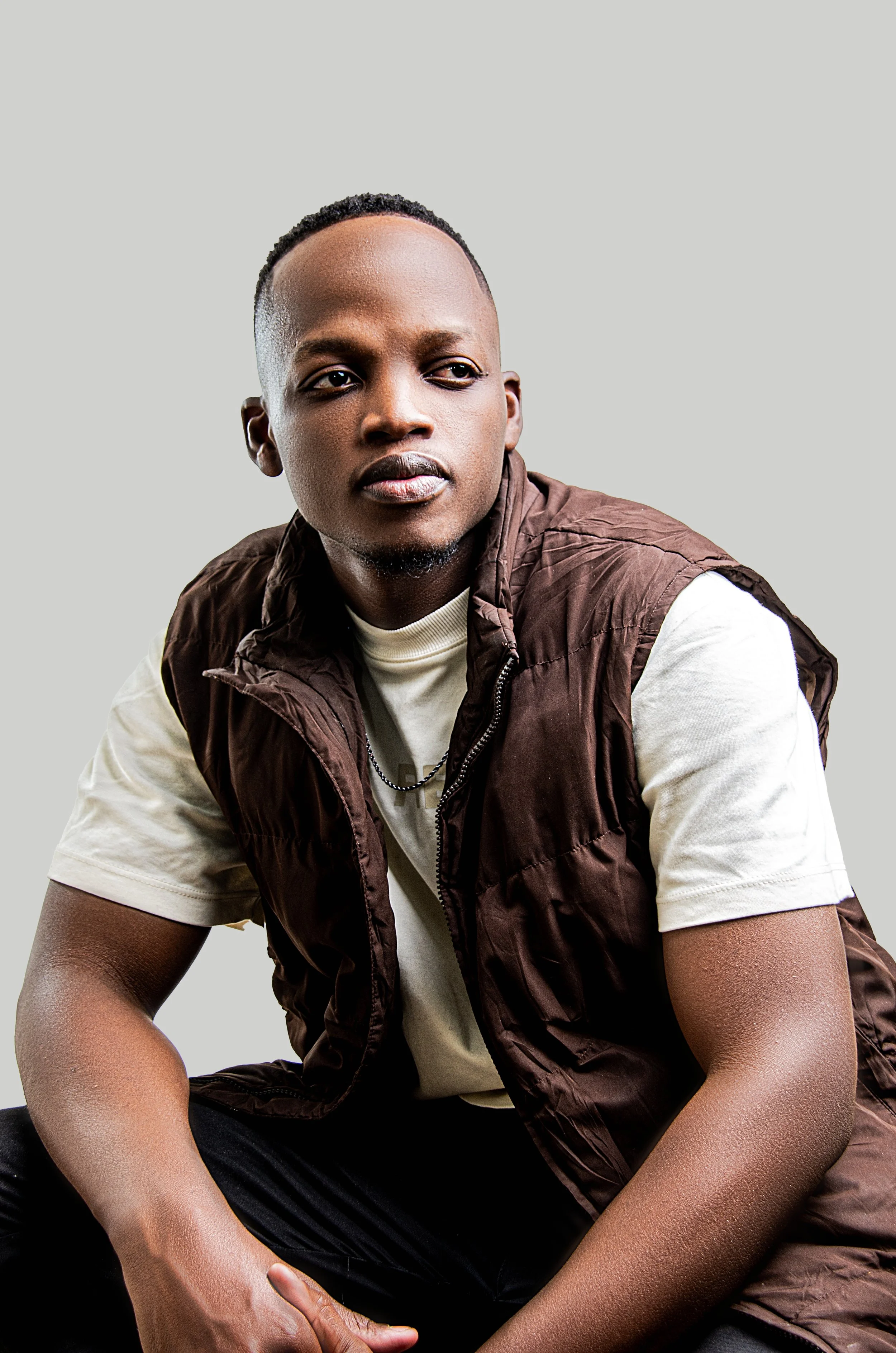 Portrait of a young African American man with short hair, sitting and looking to his right, wearing a white T-shirt and a brown puffer vest against a light gray background.