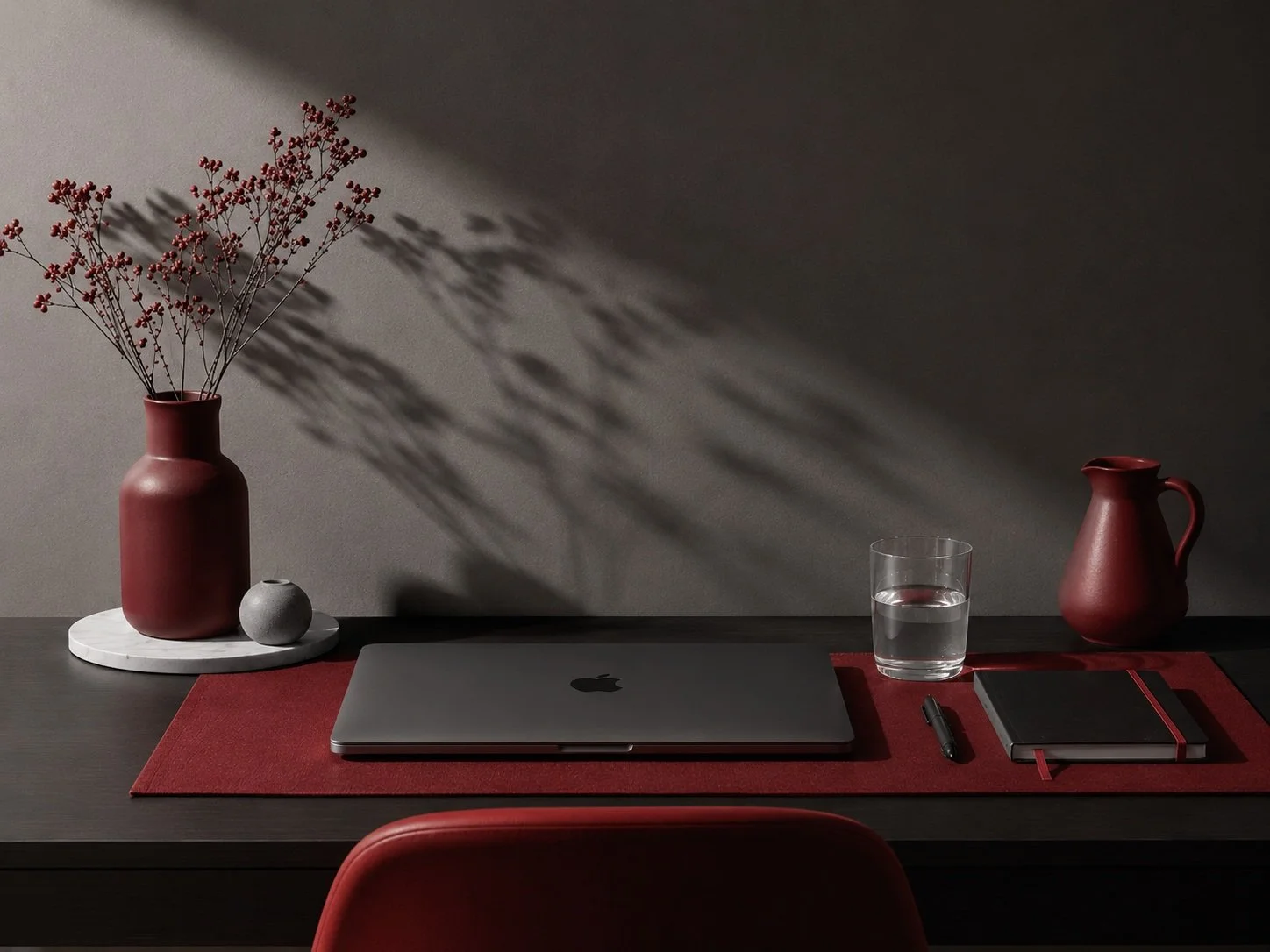 A minimalist workspace with a closed MacBook on a red desk mat, a glass of water, a black pen, a small black notebook, a red pitcher, and a red vase with dried flowers on a white circular base, against a dark gray wall with shadows.
