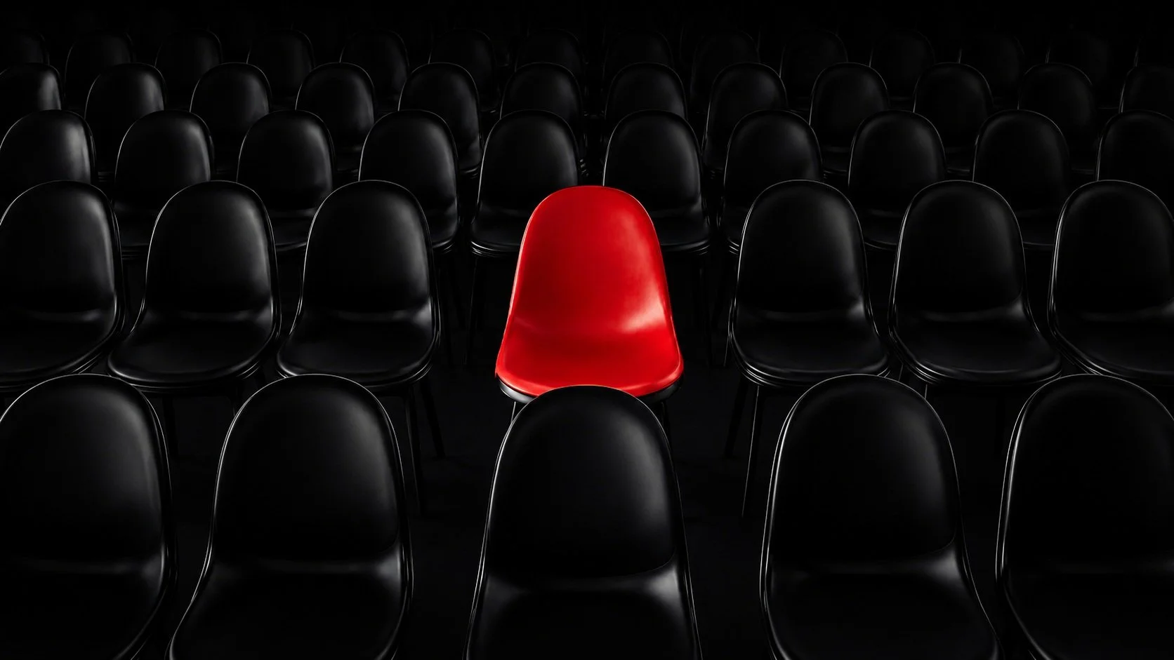 A red chair stands out among many black chairs in a dark room, with all chairs arranged in rows.