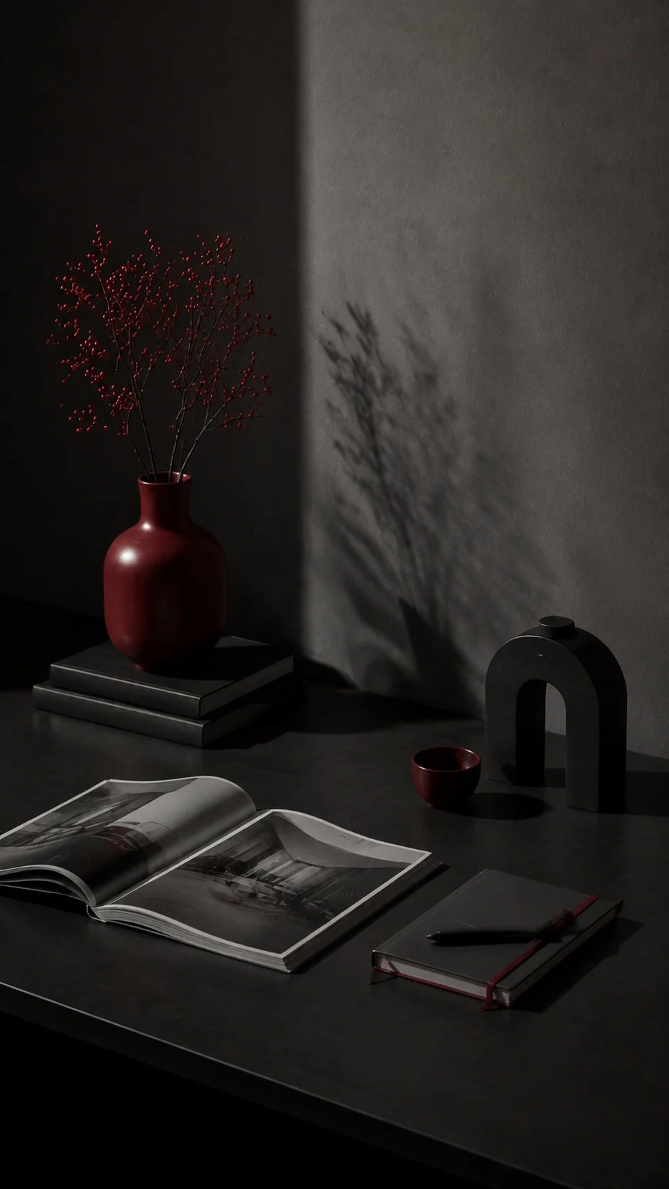 A dark desk scene with a red vase containing branches, an open book with black and white photos, a closed book with a pen on top, a small red bowl, and a black decorative object casting a shadow on the wall behind.
