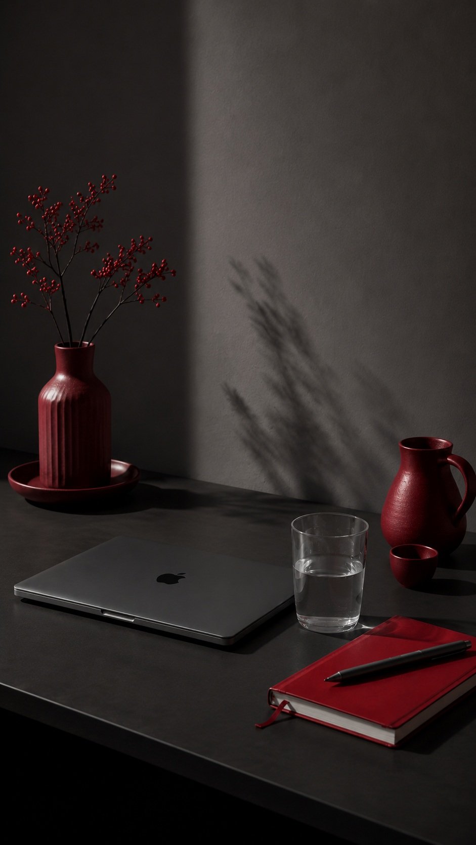 A minimalist workspace with a closed silver laptop, a glass of water, a red notebook with a black pen, and red vases with decorative branches casting shadows on a gray wall.