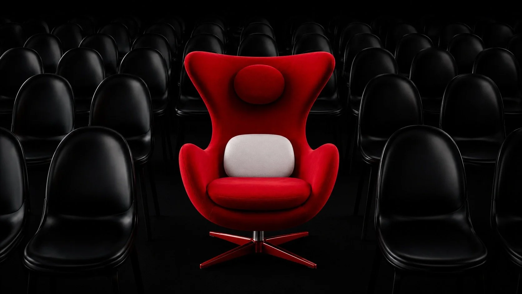 A red, high-backed, modern armchair with a white pillow in the center, surrounded by black chairs arranged in rows, in a dark setting.
