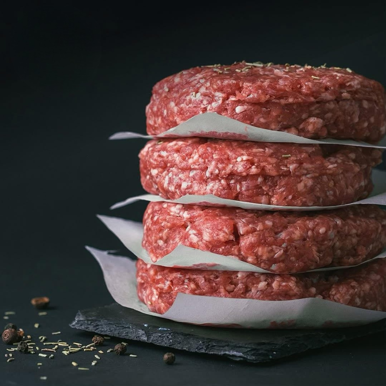 Stacked raw beef patties with seasoning on a dark surface with scattered black pepper and herbs.