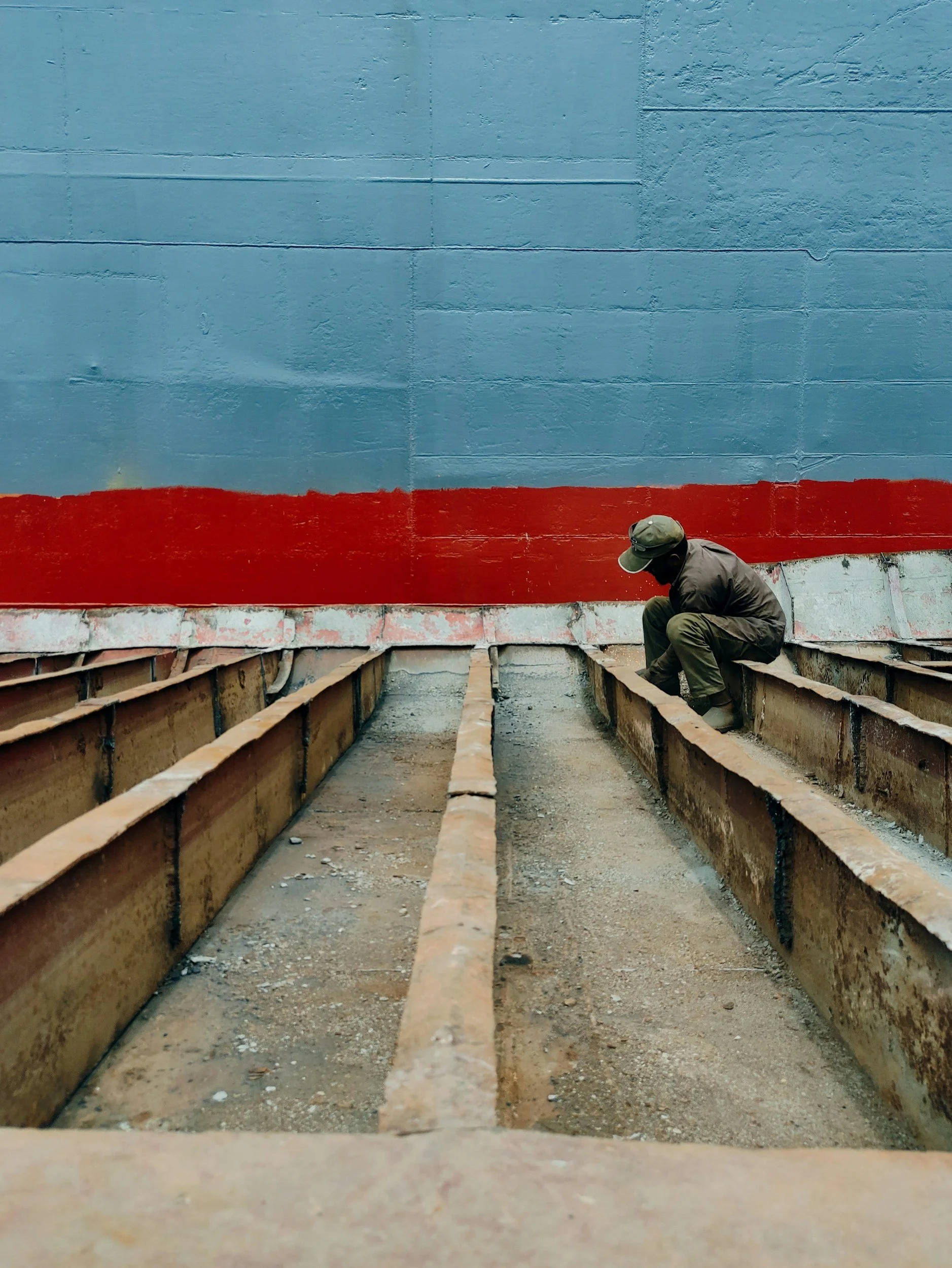 A person squat down and working on laying a foundation with wooden forms and concrete, with a painted blue wall with a red stripe in the background.
