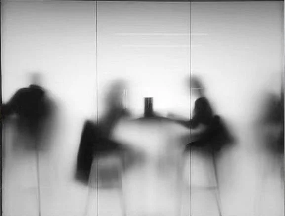 Blurred silhouettes of two people sitting at a table in front of a bright background.