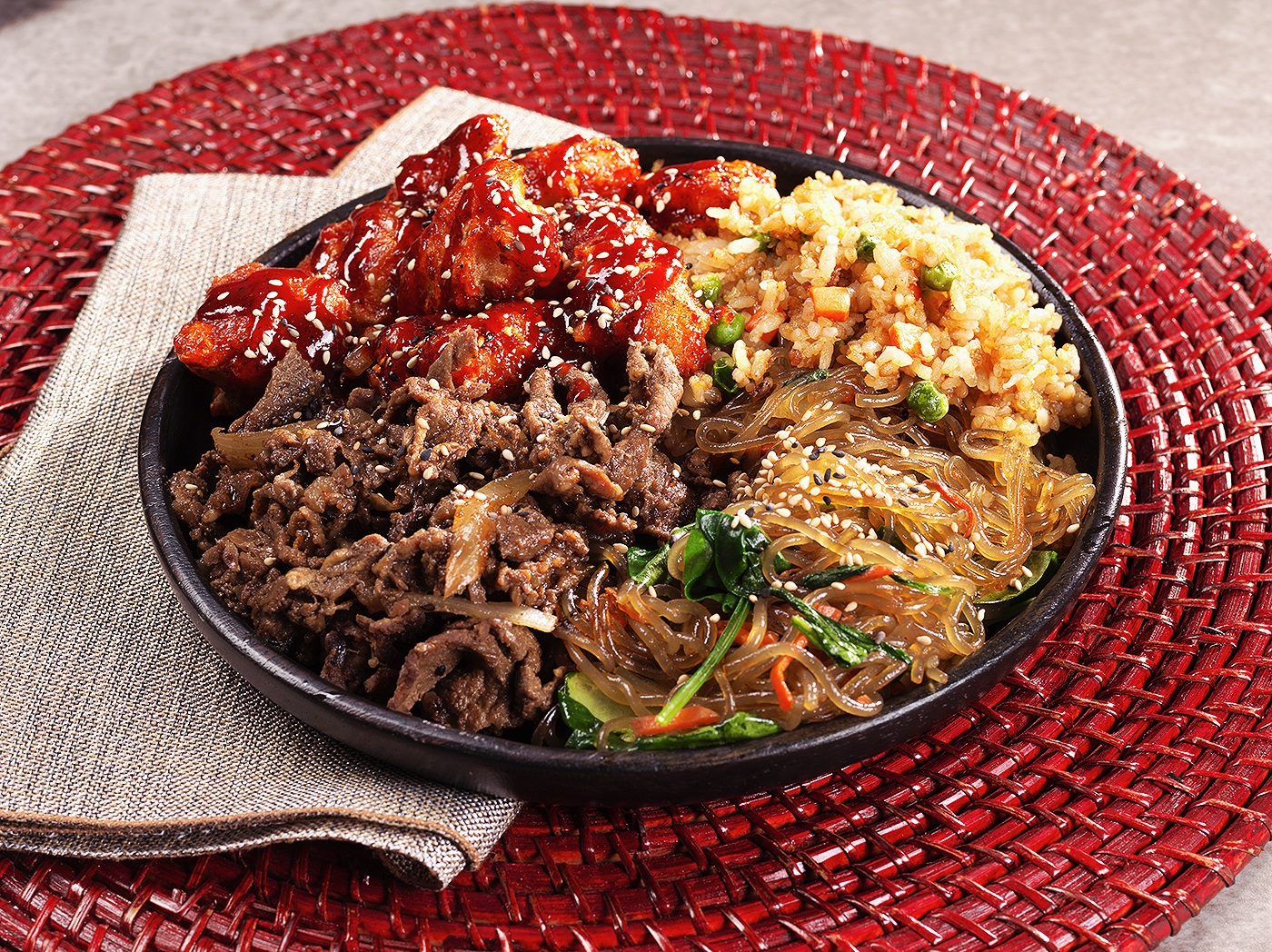 A black bowl filled with Korean dishes including spicy chicken, bulgogi beef, fried rice with peas and carrots, and stir-fried glass noodles with vegetables, all sprinkled with sesame seeds, set on a red woven placemat with a beige cloth napkin underneath.