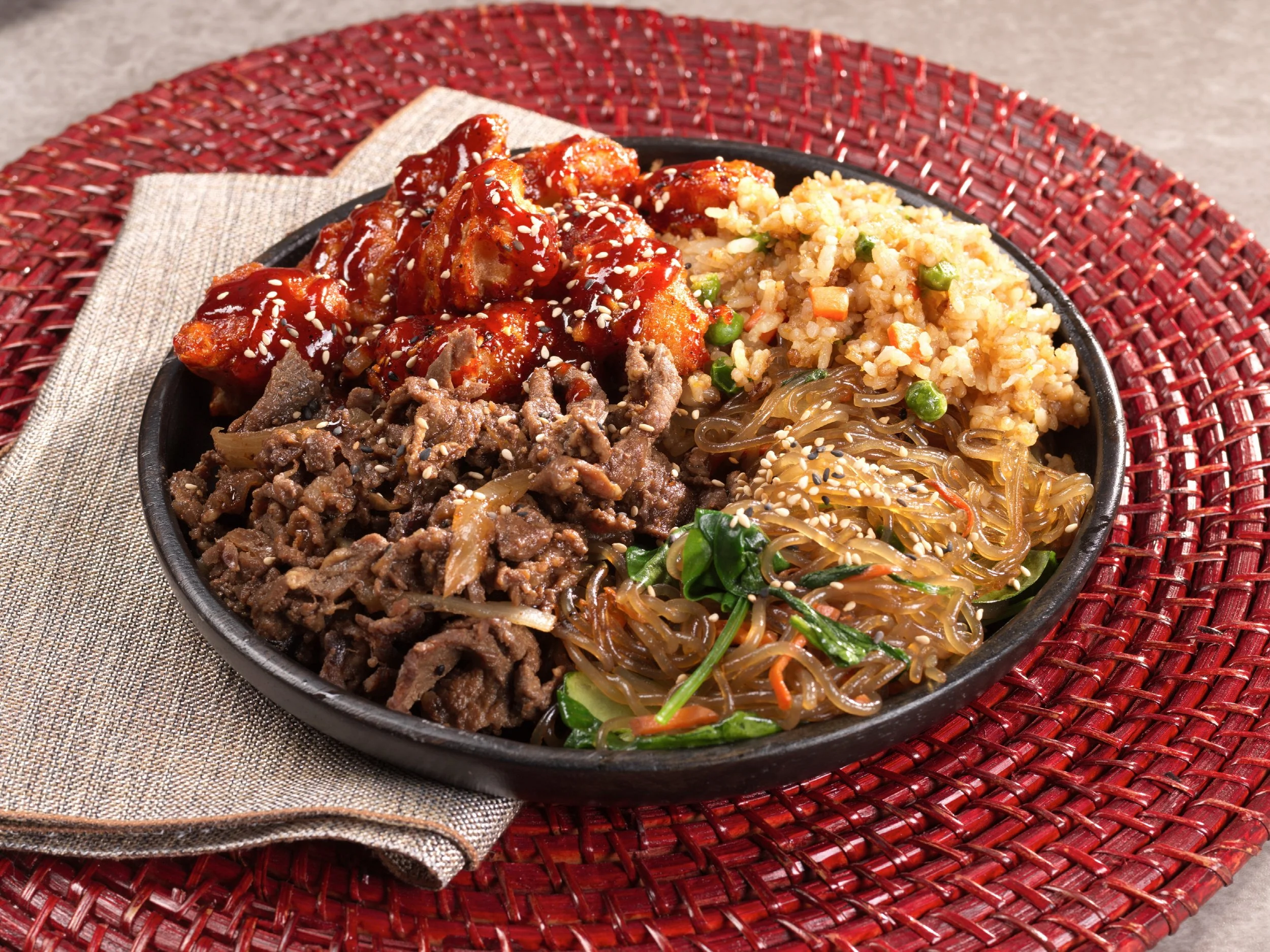 A black bowl filled with Korean dishes including spicy chicken, bulgogi, japchae noodles, fried rice with vegetables, and marinated beef, placed on a red woven placemat with a beige napkin underneath.