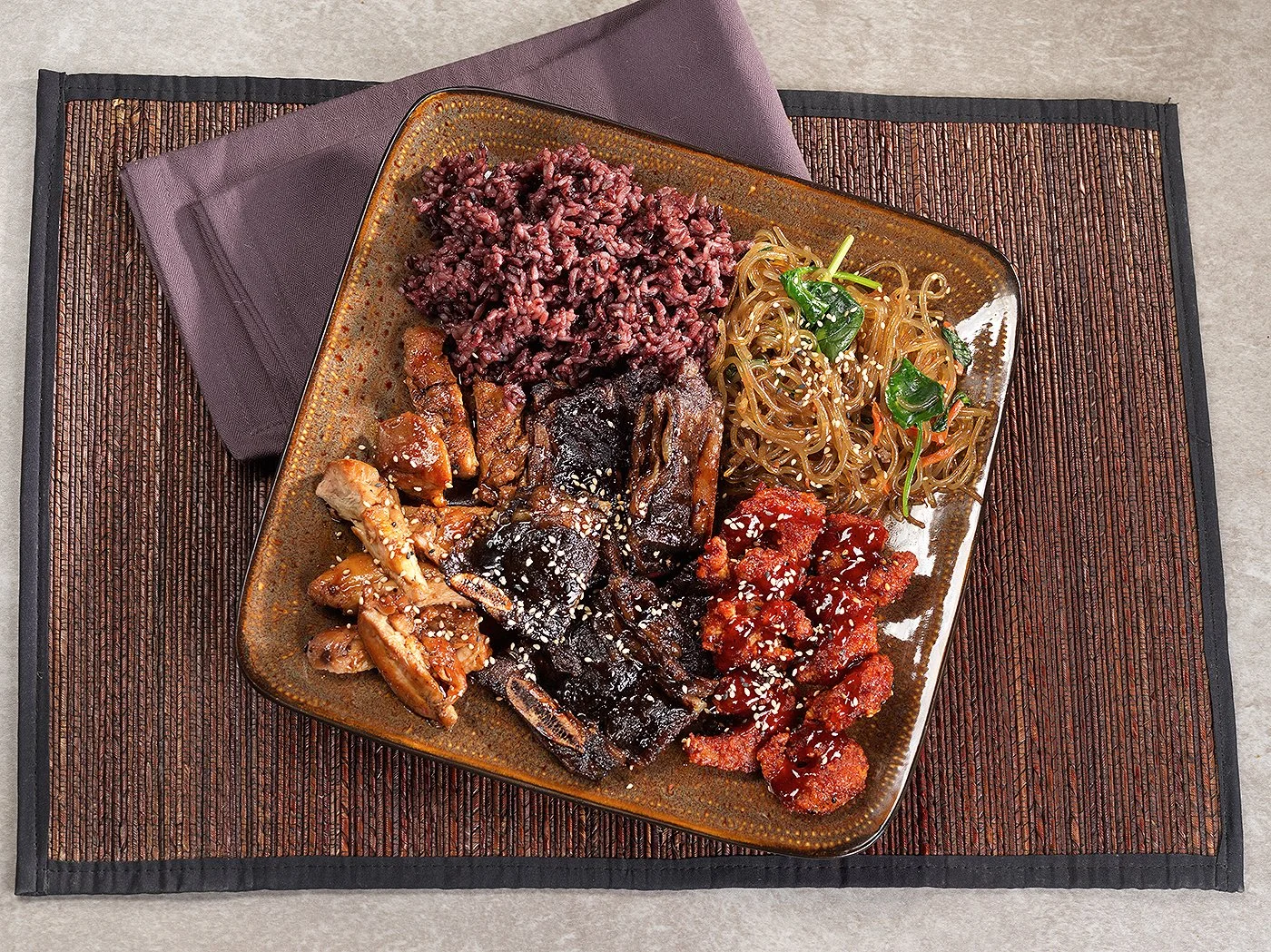 A square plate with assorted Korean dishes including rice, noodles, grilled meat, and spicy stir-fried chicken, garnished with sesame seeds.