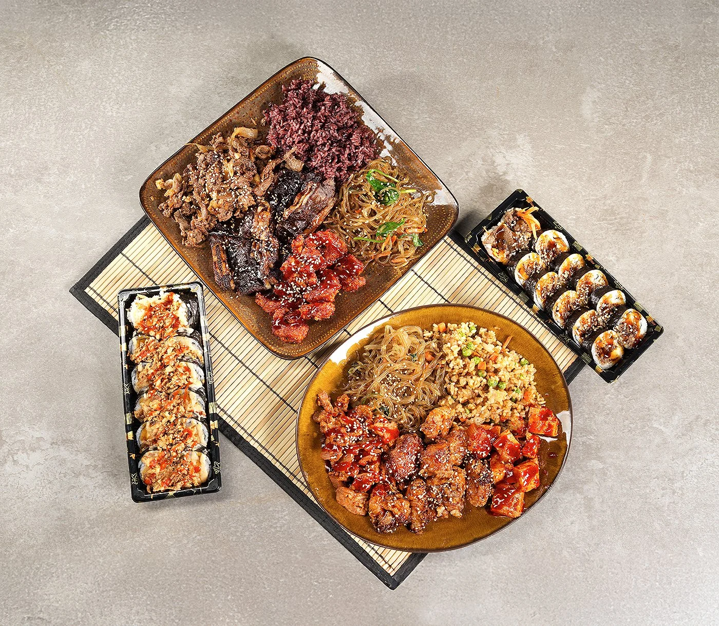 Assorted Asian dishes on a gray table, including sushi rolls, fried rice, noodles, and various meat dishes.