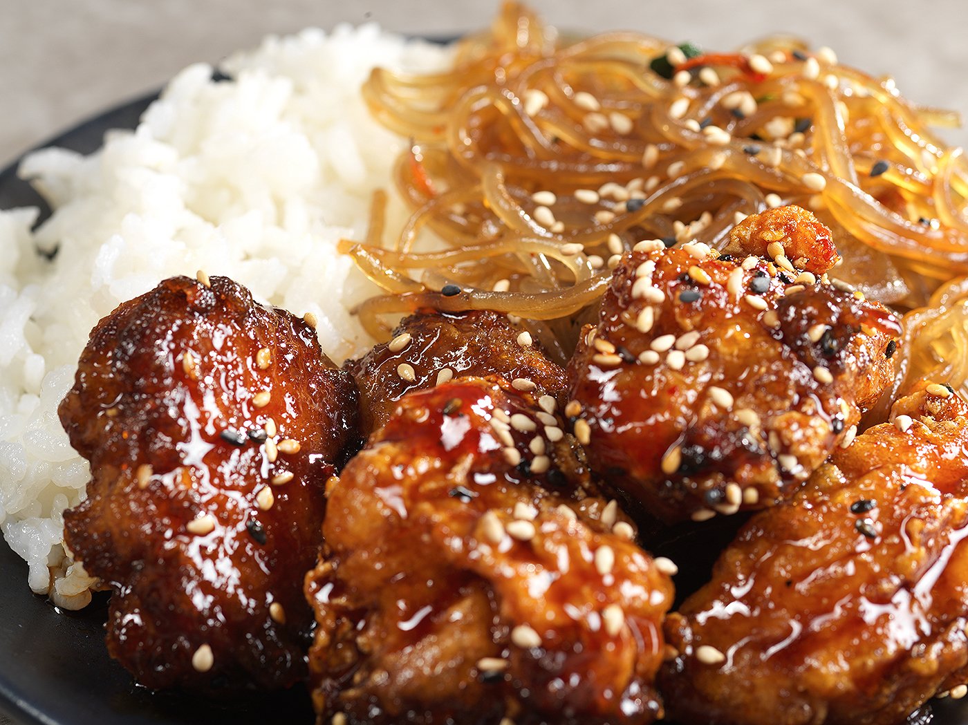 Close-up of a plate with glazed chicken wings topped with sesame seeds, white rice, and stir-fried noodles with vegetables.