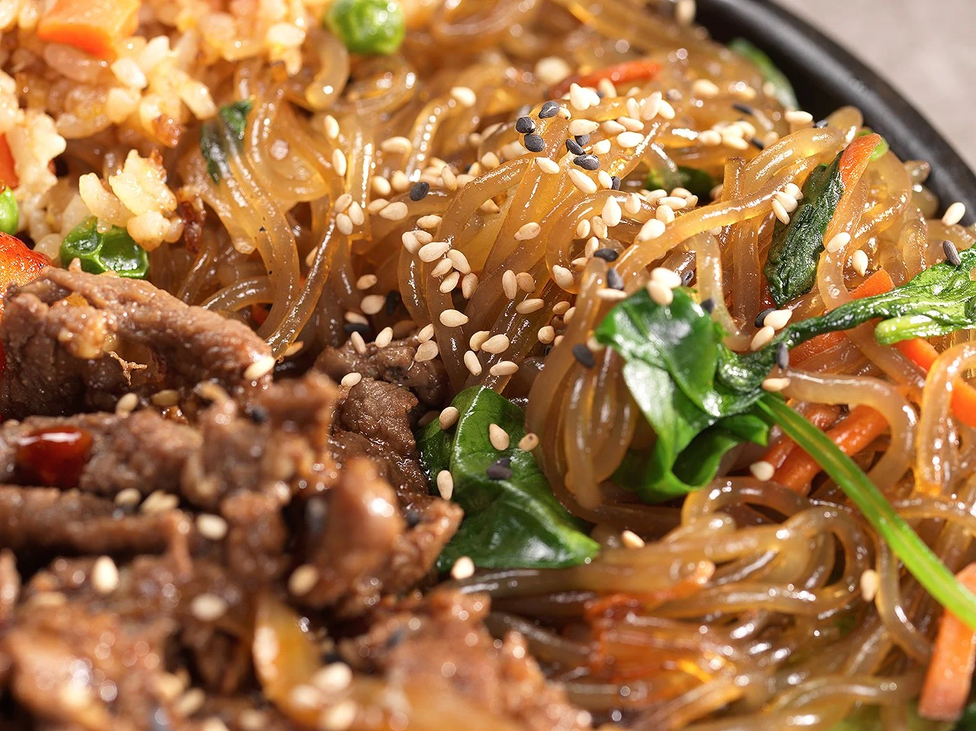 Close-up of a plate of stir-fried noodles with vegetables and sesame seeds, featuring bean sprouts, spinach, and shredded carrots, with beef on the side.