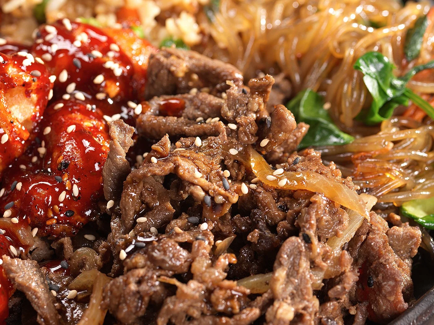 Close-up of a plate with beef, noodles, and vegetables, garnished with sesame seeds, topped with red sauce.