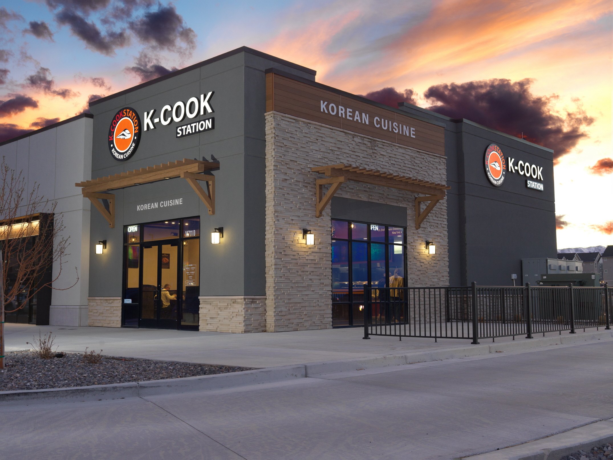 A modern commercial building with signs for K-Cook Station and Korean Cuisine, featuring large windows, decorative wooden awnings, exterior lighting, and a parking lot in front, with a sunset sky in the background.