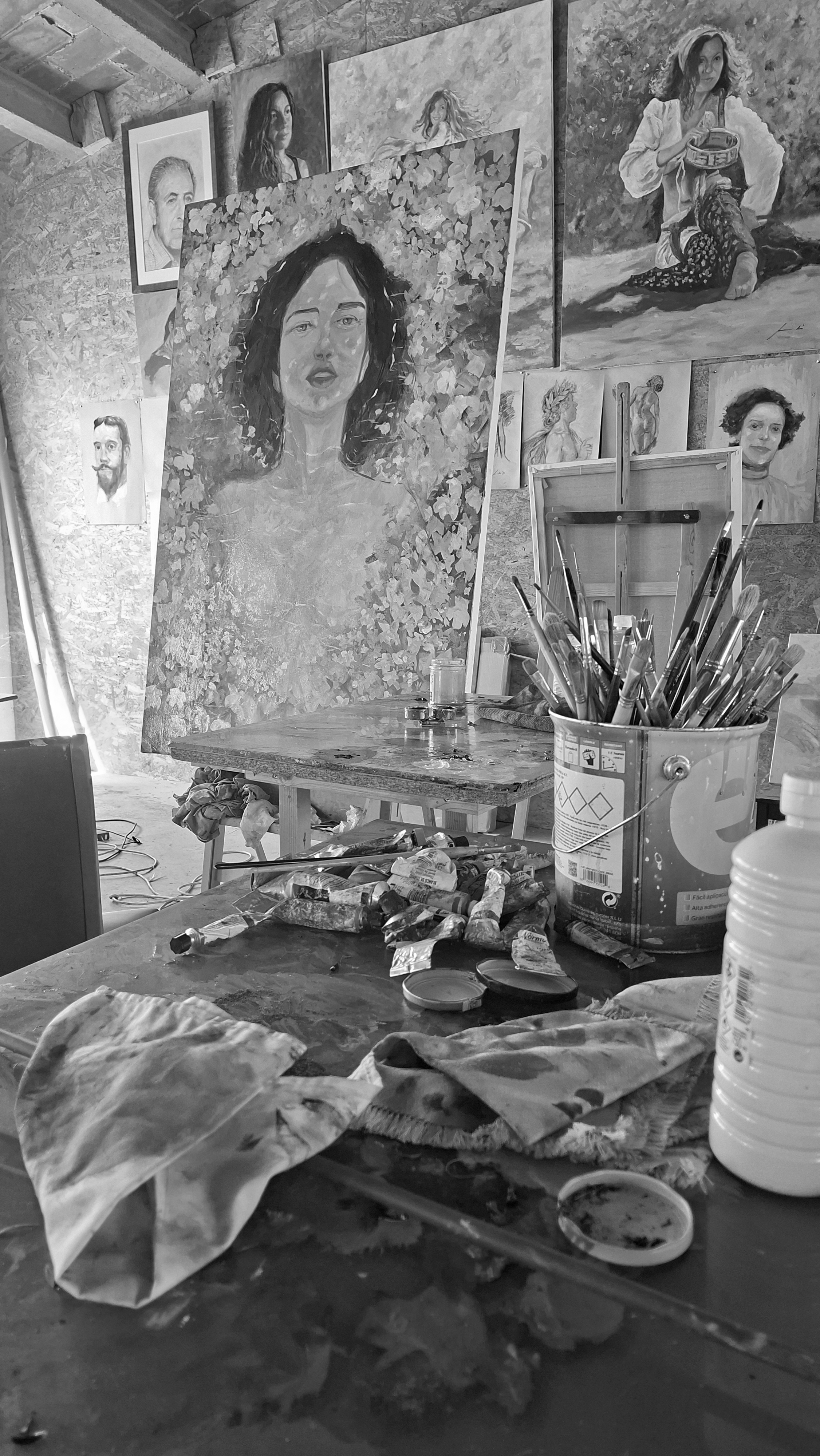 Estudio de arte con varias pinturas en las paredes y en caballetes, incluyendo retratos y pinturas de mujeres. En primer plano hay una mesa con tubos de pintura, pinceles, una taza con agua, y materiales de pintura. El ambiente parece un taller de artista con paredes de piedra y techo de madera.