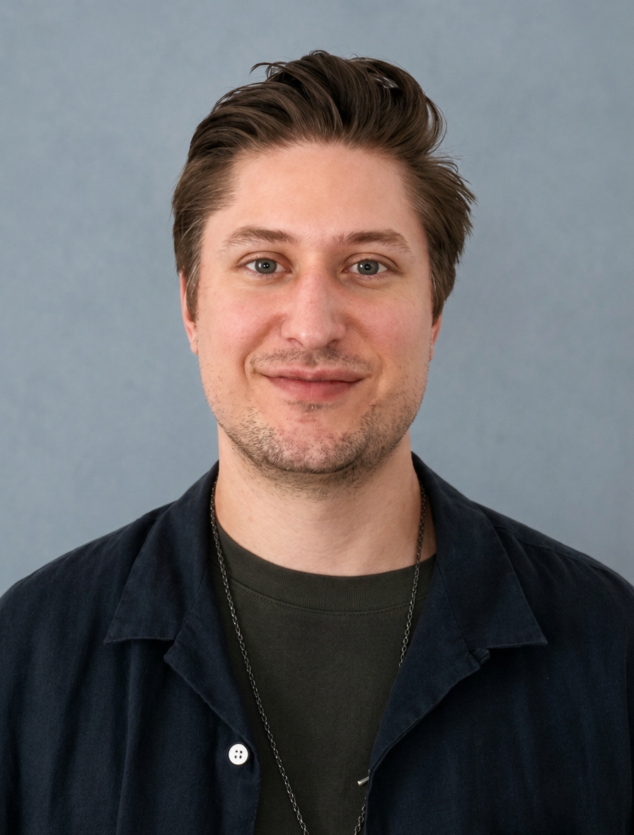 Portrait of a young man with brown hair, blue eyes, and a light beard, wearing a black shirt and a dark jacket, standing against a plain gray background.
