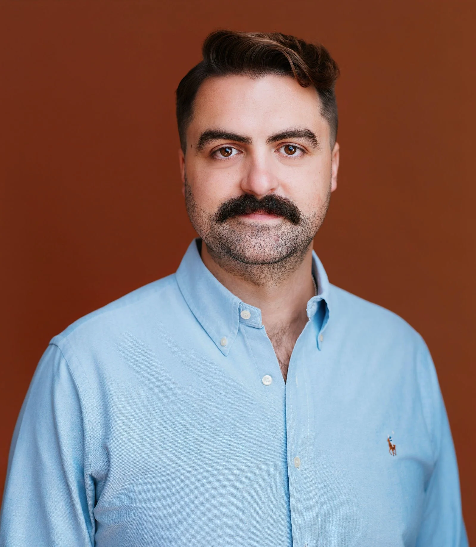 A man with dark hair, a mustache, and light skin wearing a light blue button-up shirt with a small embroidered logo on the left side of the chest, standing against a solid brown background.