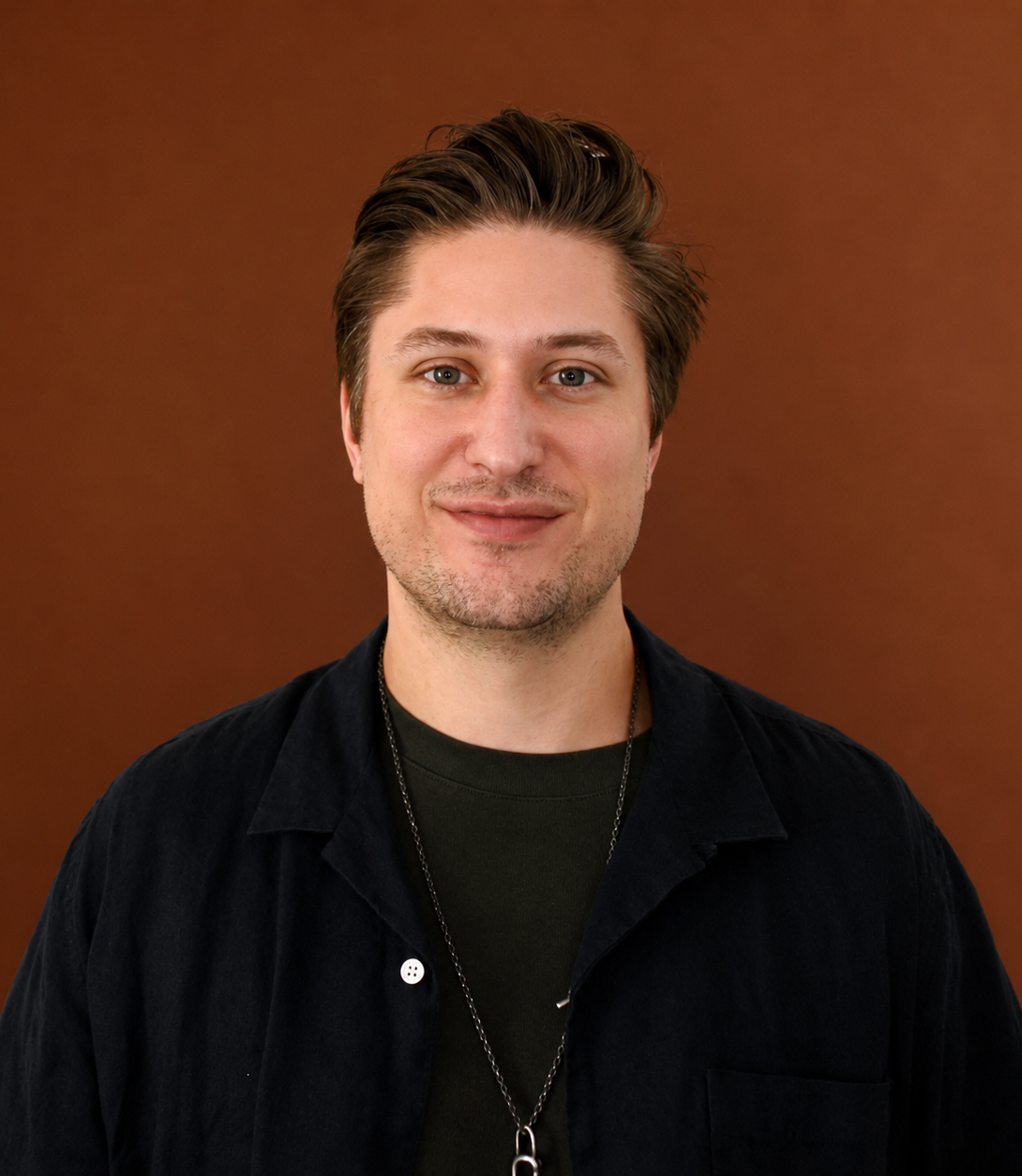 A young man with short brown hair, fair skin, and light facial hair, wearing a black shirt and a silver chain necklace, stands against a brown background looking at the camera and smiling gently.