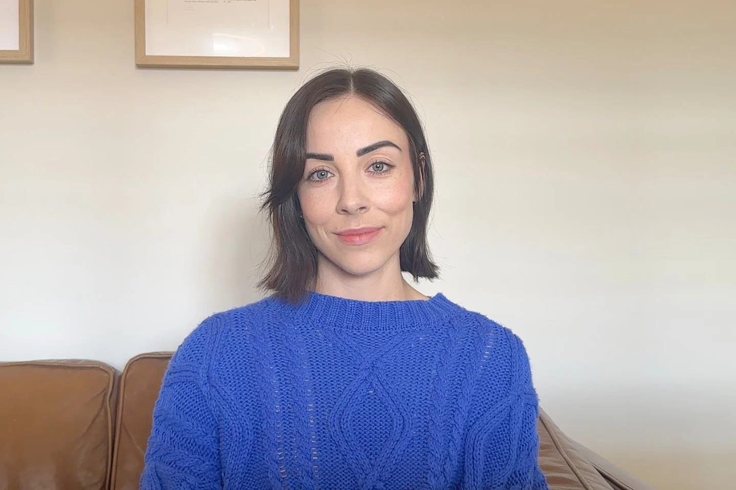 A woman with short dark hair and blue eyes wearing a bright blue knit sweater, sitting on a brown leather couch with a neutral wall and framed pictures behind her.