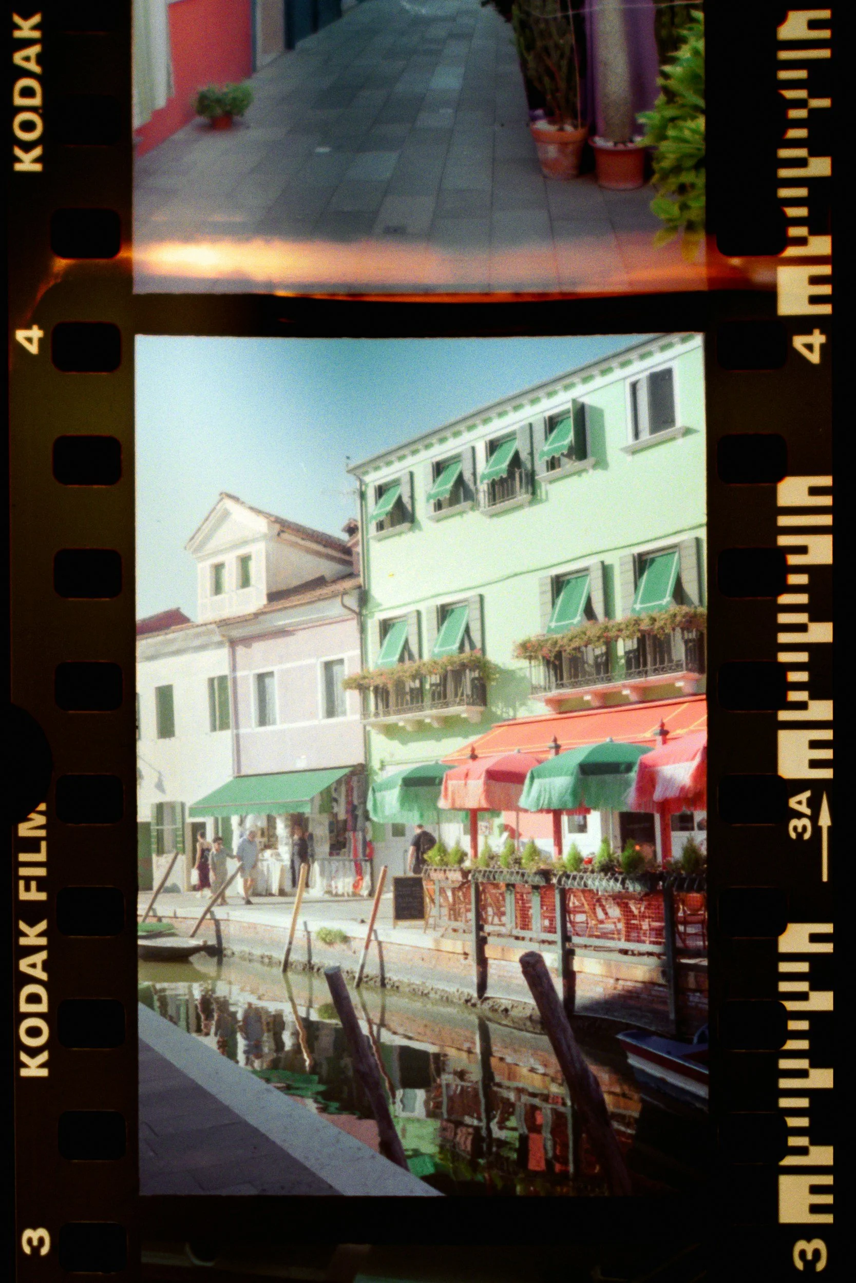 View of a waterfront town street with pastel buildings, outdoor cafes with colorful umbrellas, and a canal reflecting the buildings.
