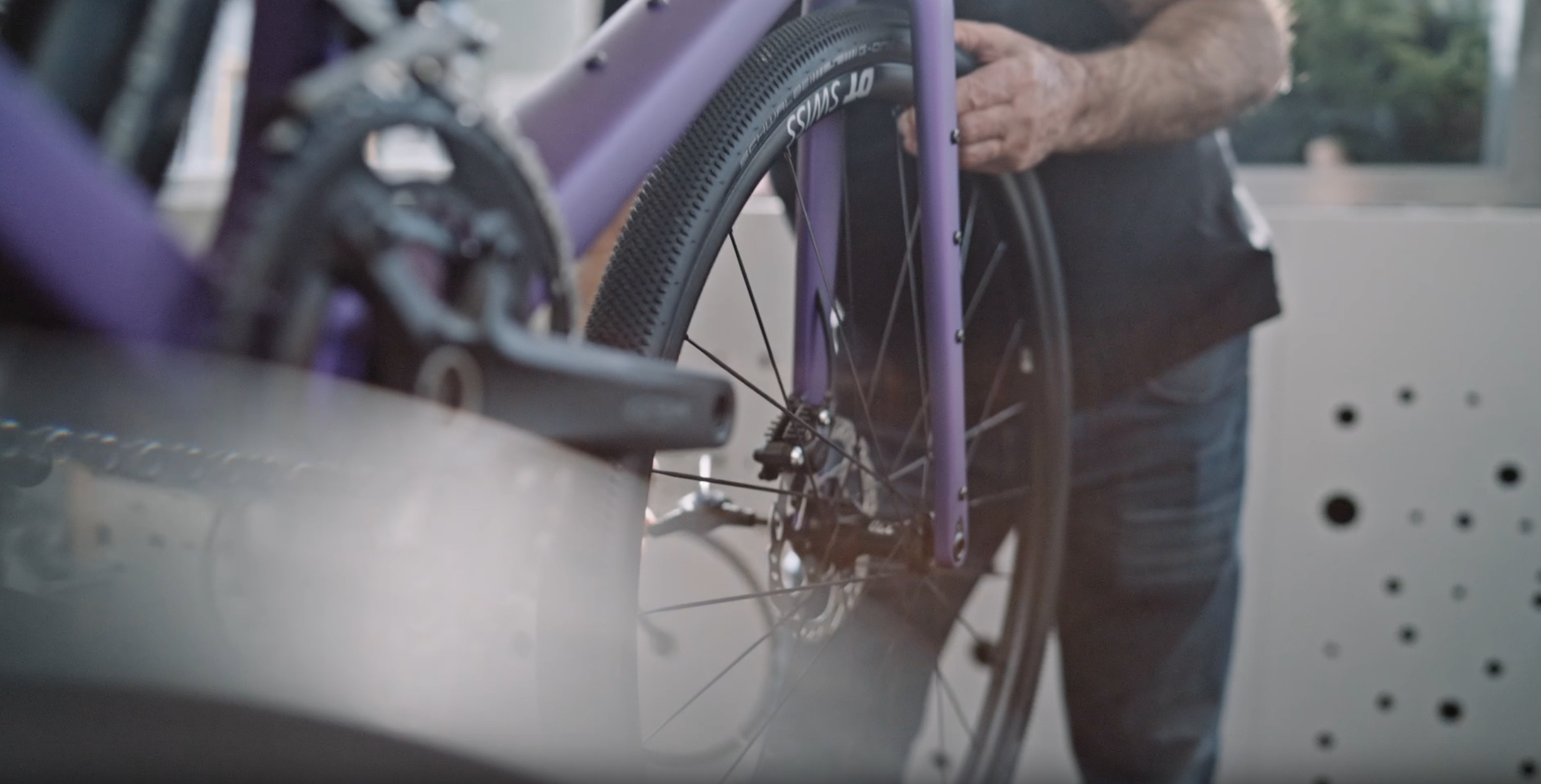 Nahaufnahme eines Fahrrads mit purple Rahmen, Fahrradkette und Reifen, im Hintergrund eine Person in dunkler Kleidung.