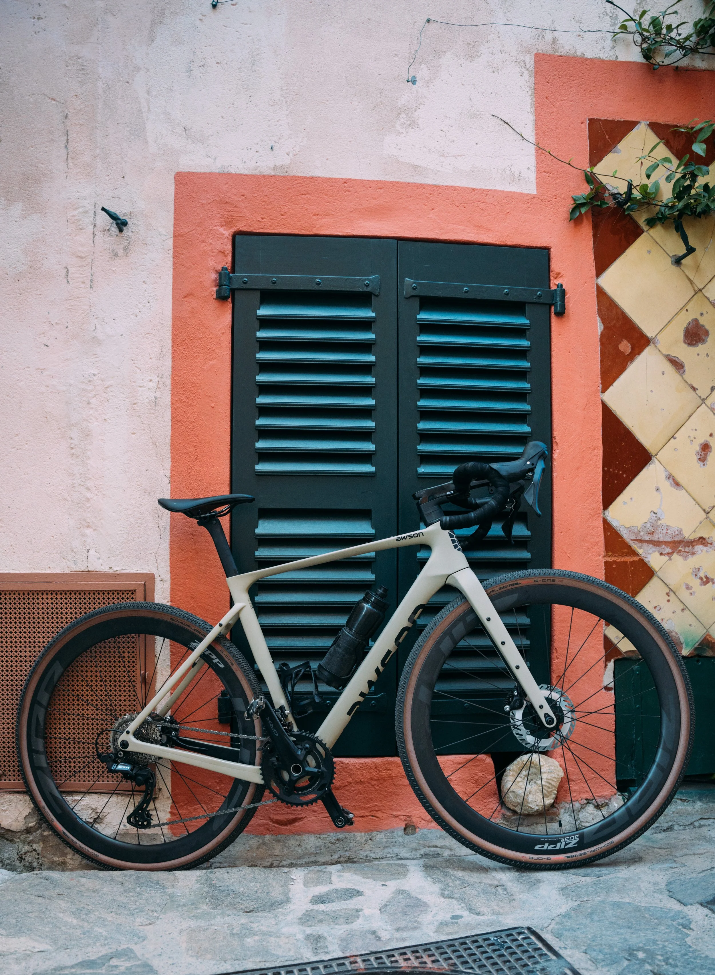 Ein weißes Fahrrad mit dunklen Reifen steht vor einem bunten Haus mit orangenen und gelben Fliesen und einer schwarzen Tür mit Lüftungsschlitzen, im Hintergrund eine Hauswand mit einem Fenster und Pflanze.
