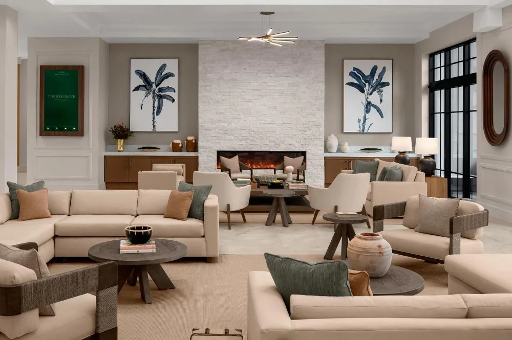 Modern living room with beige sofas, armchairs, a central fireplace with a white brick wall, abstract artwork, and large windows.