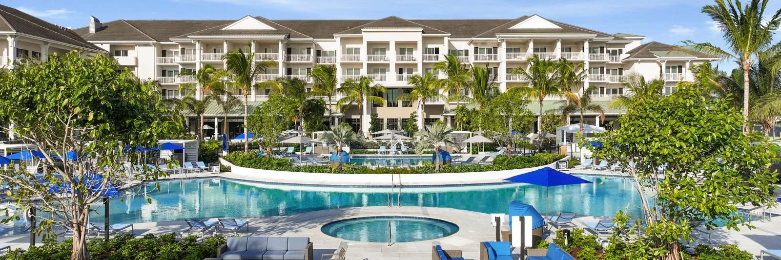 View of a large luxury hotel with a swimming pool and hot tub, surrounded by palm trees and lounge chairs, with a multi-story building in the background.