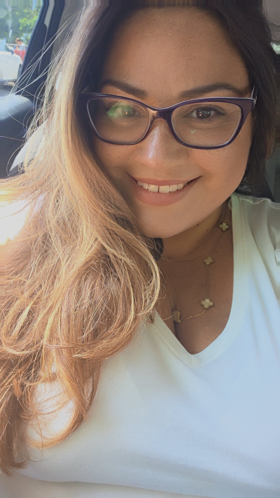 Smiling woman with long, wavy, light brown hair, wearing glasses, a white top, and gold jewelry, sitting in a car.