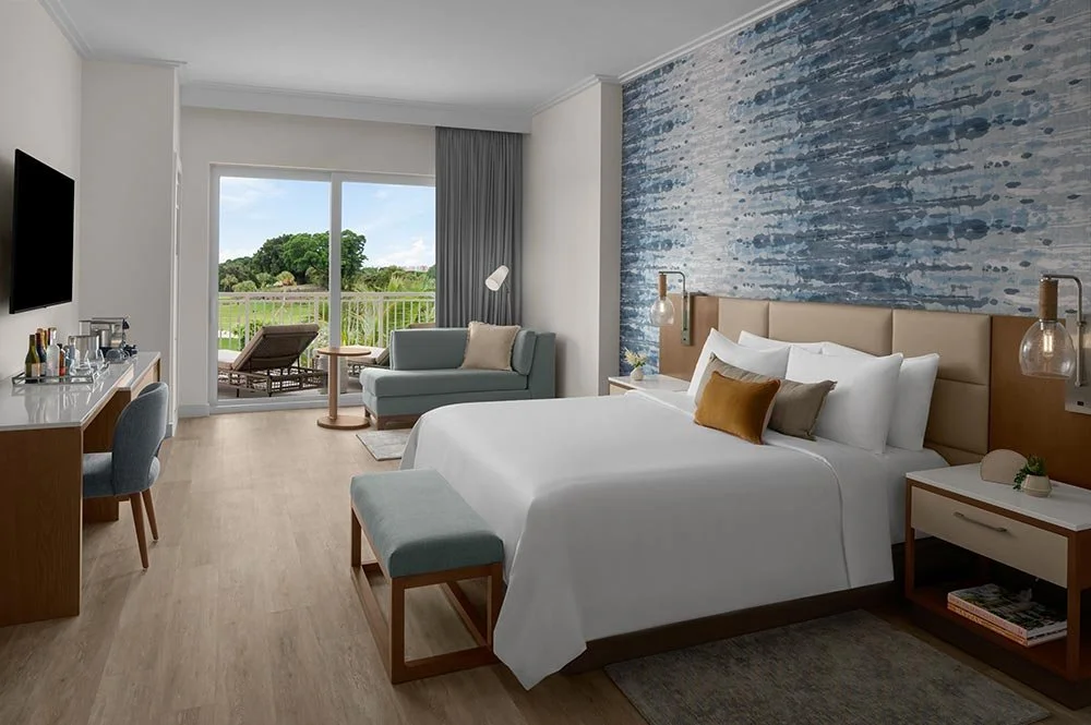 A modern hotel room with a large bed, blue accent wall, small sofa, balcony with outdoor seating, and a view of greenery outside.