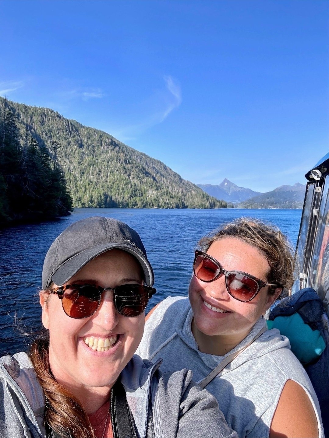 Two women on a boat smiling with sunglasses, water, mountains, and a clear blue sky in the background.