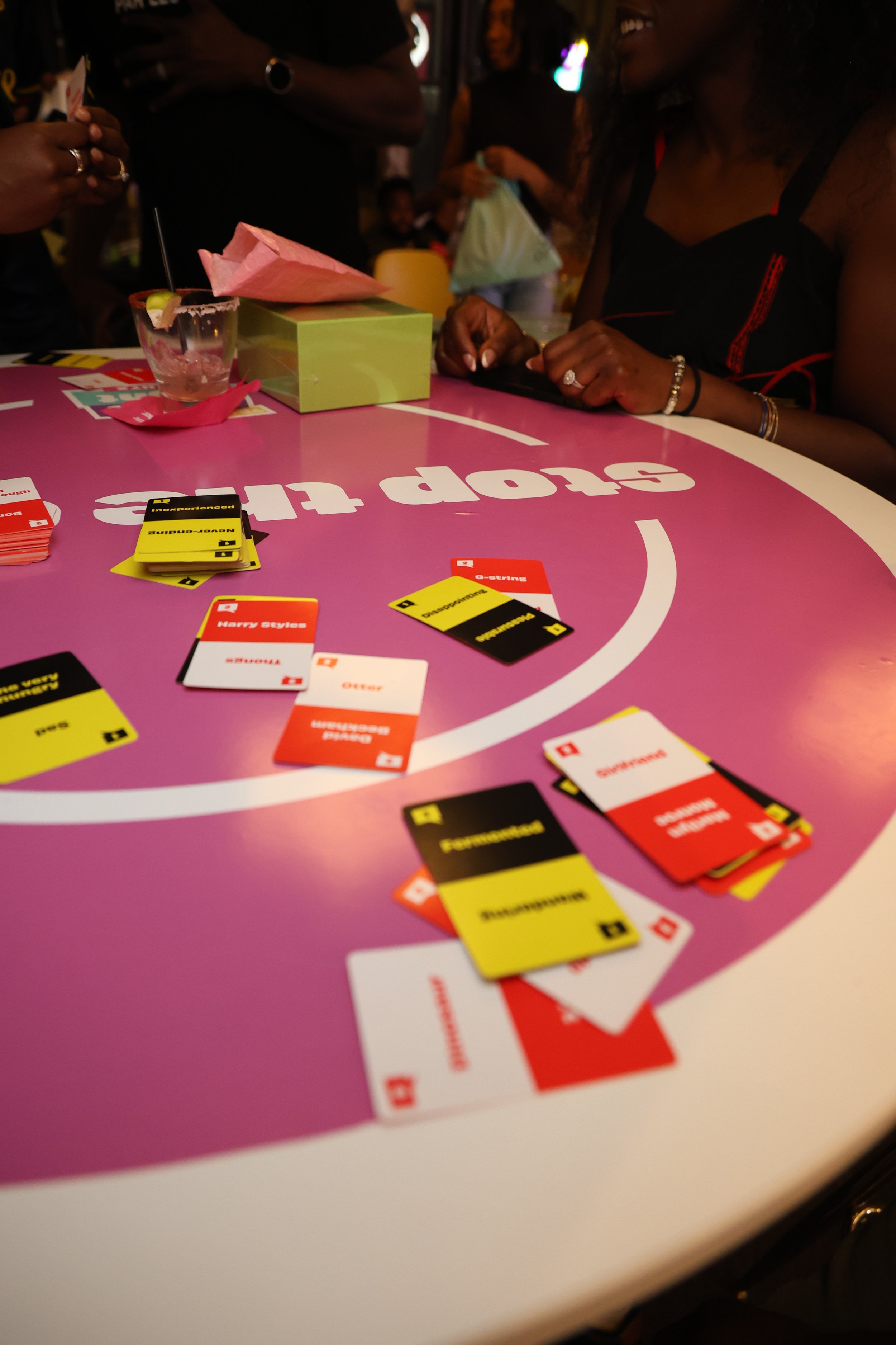 People playing a card game at a pink table in a lively setting. Cards are spread out on the table with some face up. The individuals are smiling and appear to be enjoying themselves, with colorful lighting in the background.