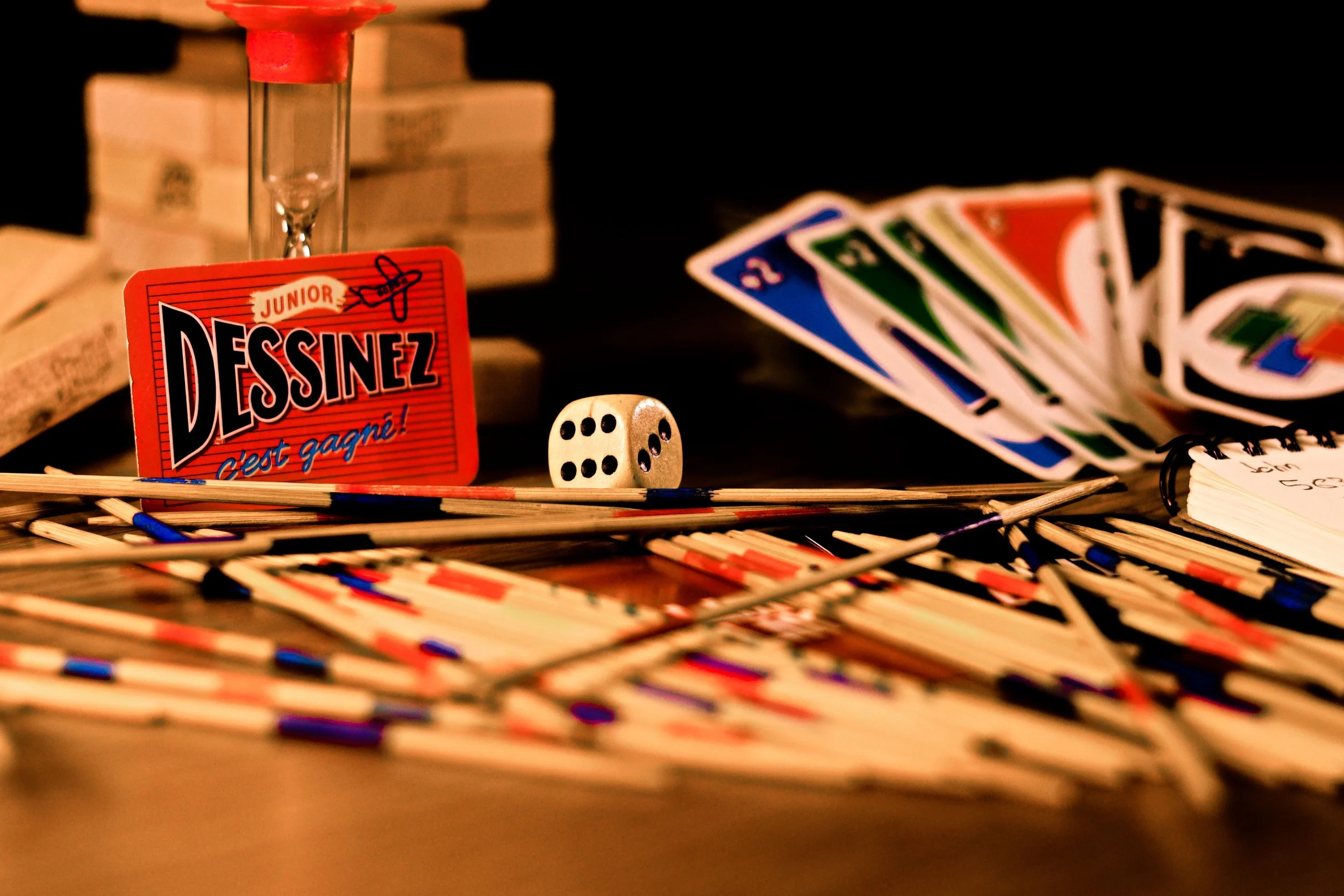 A tabletop board game setup with miniatures, cards in a fan, a dice, and a red 'Dessinez, c'est gagné!' sign with a signature, against a background of stacked boxes.