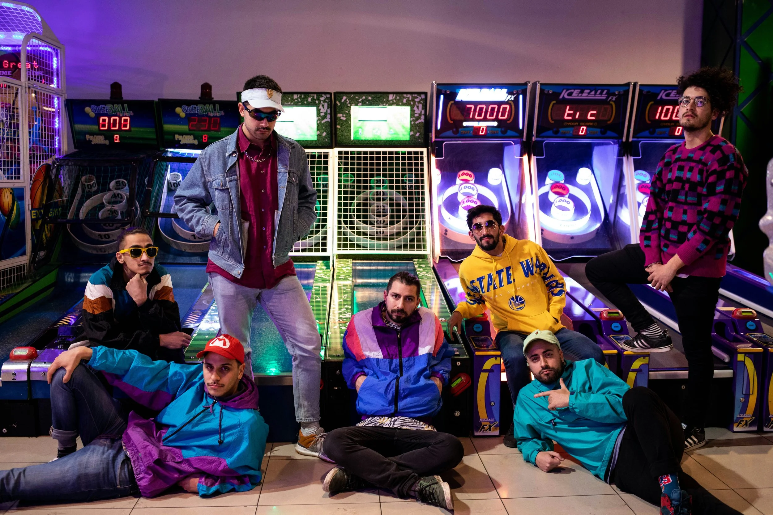 Group of seven young people in colorful retro and sporty clothing posing in front of arcade skee-ball and pinball machines at an indoor arcade. Some are sitting, some are standing, with relaxed and confident expressions.
