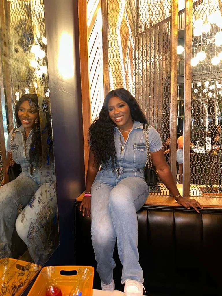 A woman with long, curly black hair, wearing a light blue denim jumpsuit, sitting on a black cushioned bench in a restaurant or bar. She is smiling, holding a black bag over her shoulder, with a pink wristband on her left wrist. There is a mirror reflecting her image, and a decorative wall with warm lighting behind her. An orange tray with a drink and a pitcher is in the foreground.