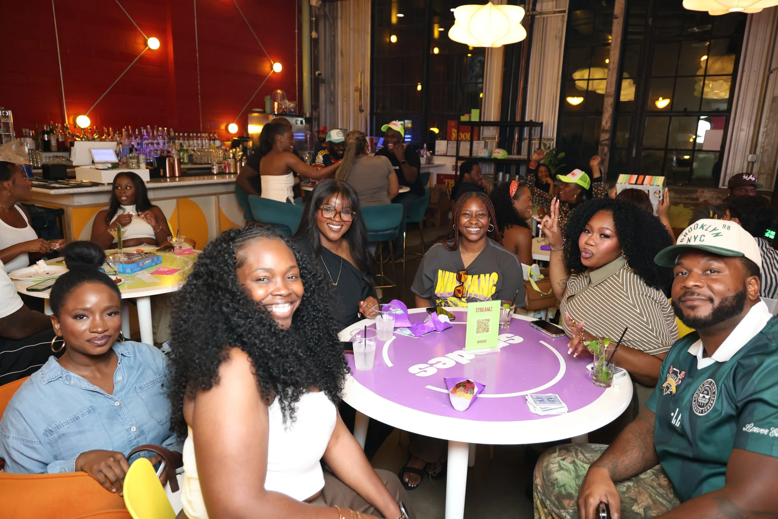 A group of smiling people gathered around a table at a party in a modern restaurant or bar with large windows and colorful decor, celebrating and enjoying drinks.