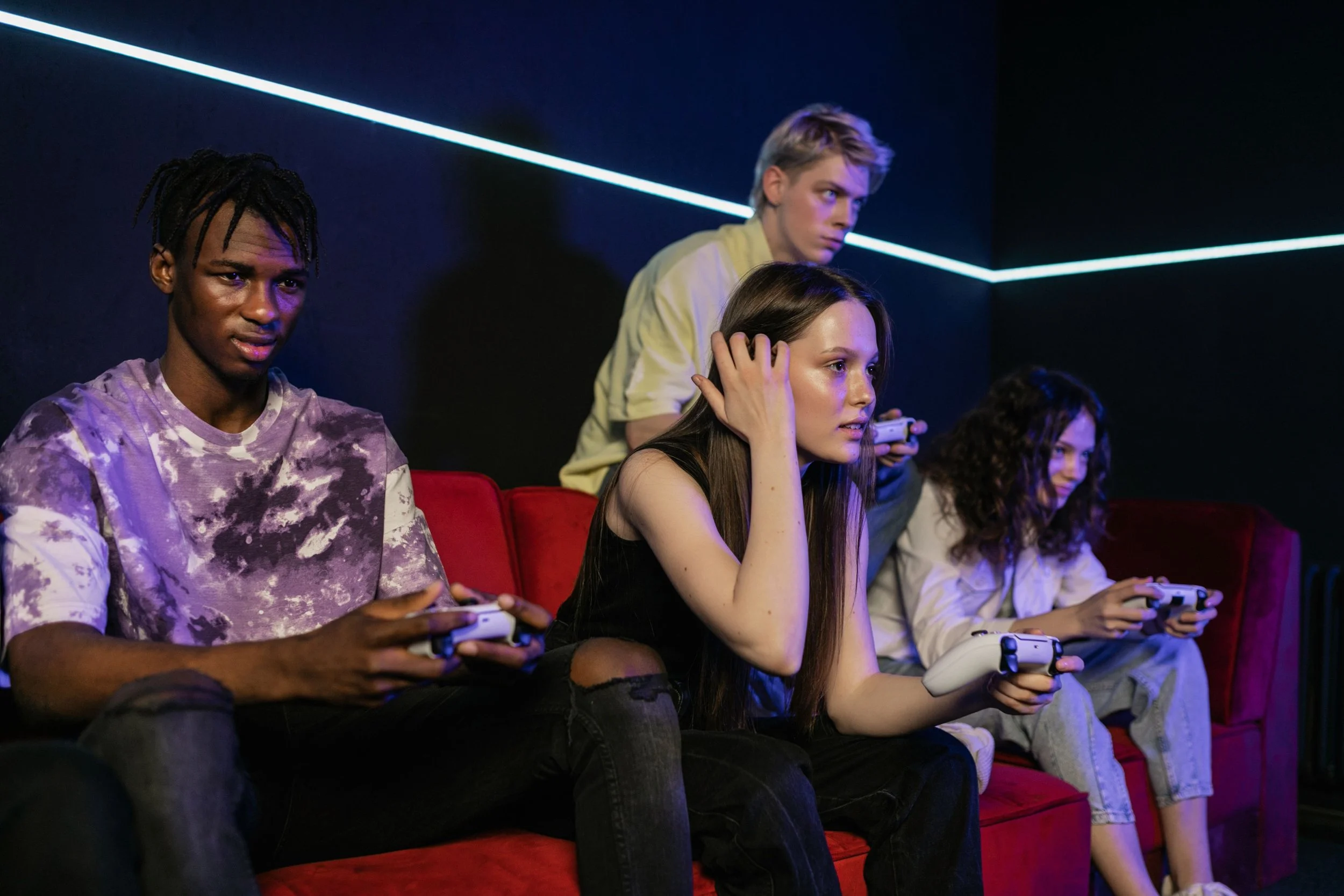 A group of young people sitting on red velvet couches in a dark room illuminated by blue LED strip lights, playing video games with controllers.