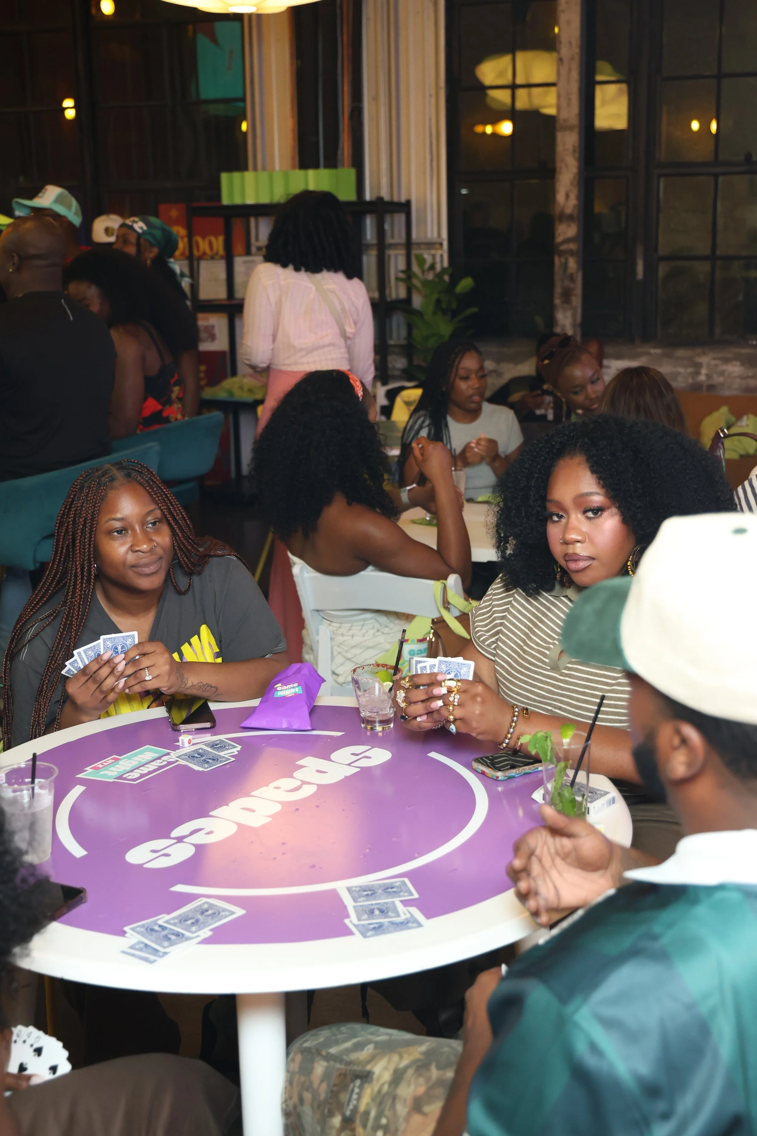 People gathered around a purple table playing cards in a lively indoor setting, with large windows and warm lighting.