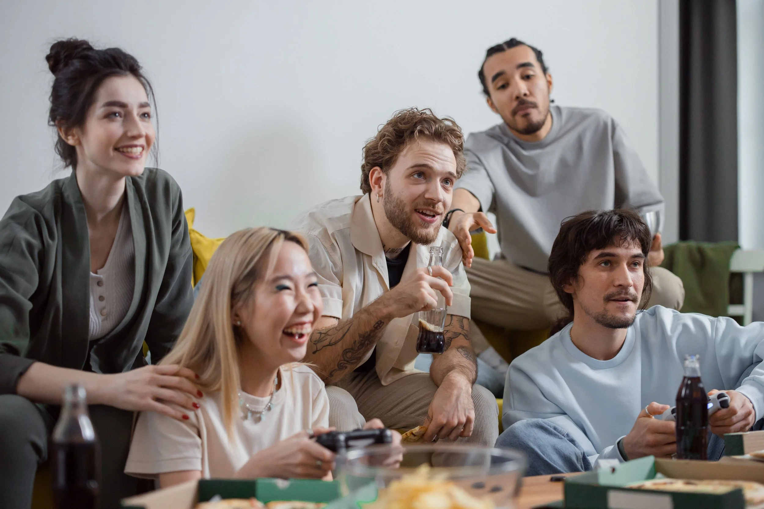 A group of young adults gathered in a room, watching something off-camera, some smiling and holding drinks or gaming controllers.