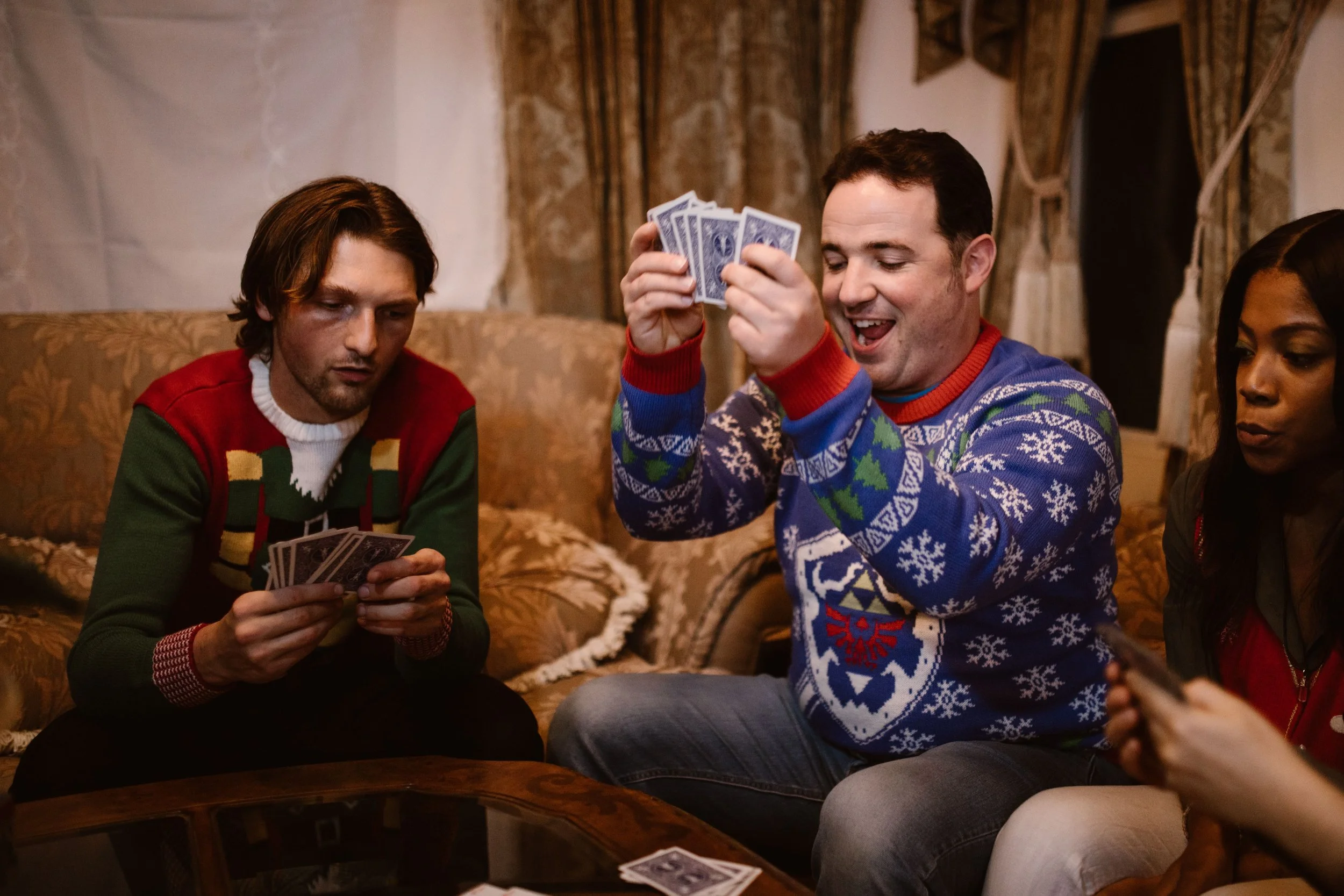Four people sitting on a sofa playing cards, three women and one man, all wearing festive sweaters, in a warmly decorated room with curtains.